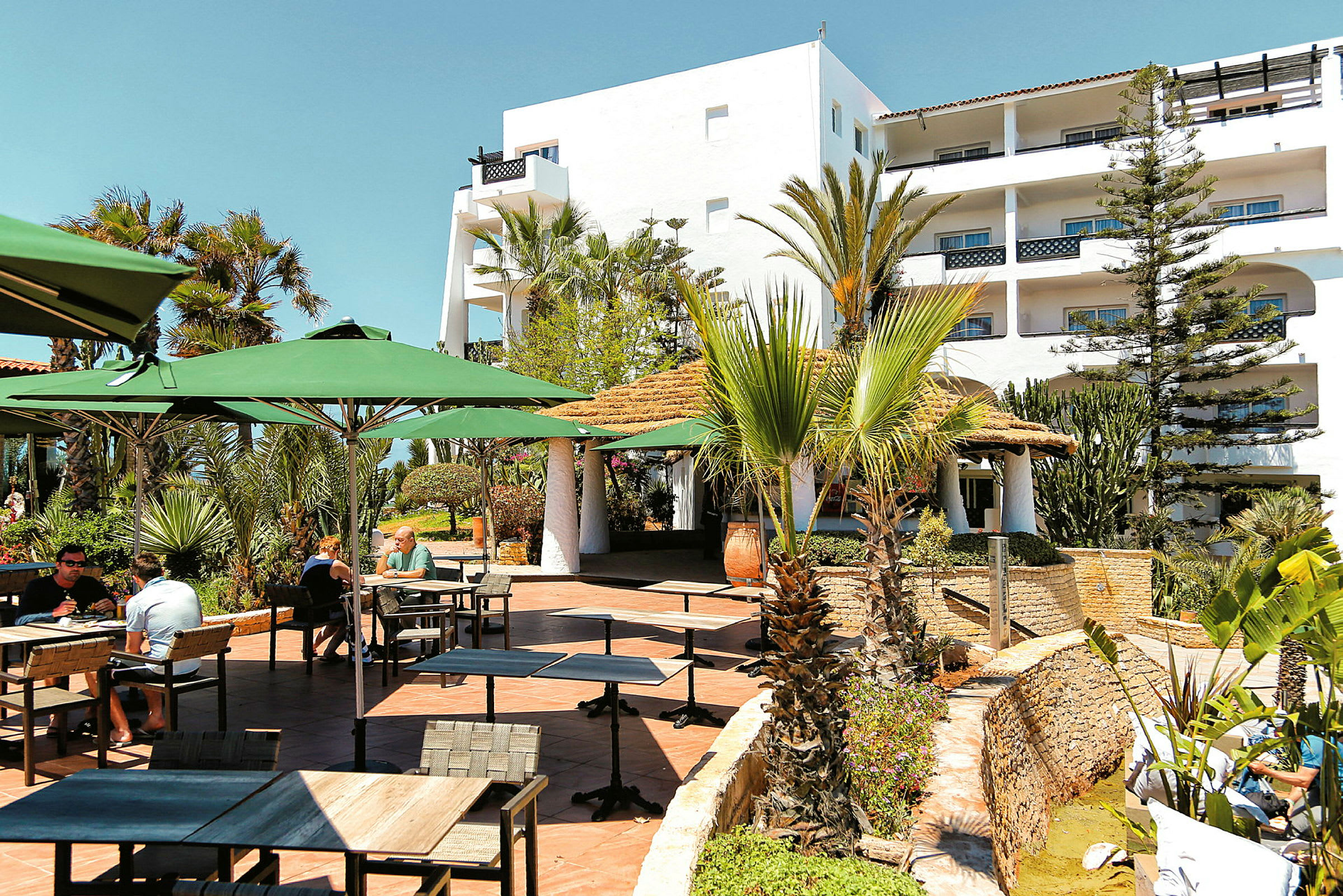 Ulkoravintola, jossa pöytiä ja tuoleja vihreiden aurinkovarjojen alla, ympärillä kasveja ja palmuja. (Tekoälyn luoma teksti)