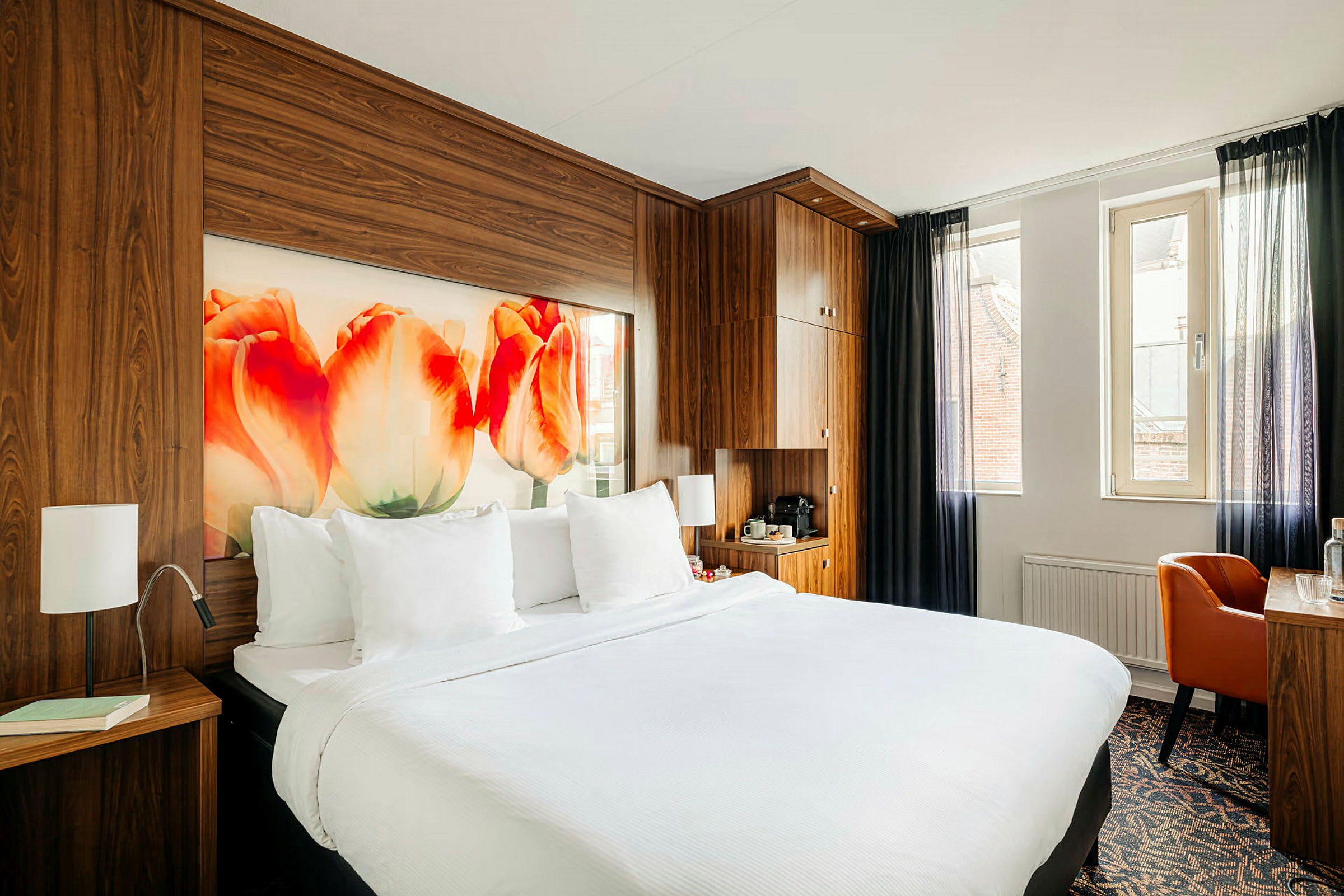 Hotellihuone, jossa parisänky, puupaneelit, tulppaanikoriste seinällä ja kirjoituspöytä ikkunan vieressä. (Tekoälyn luoma teksti)