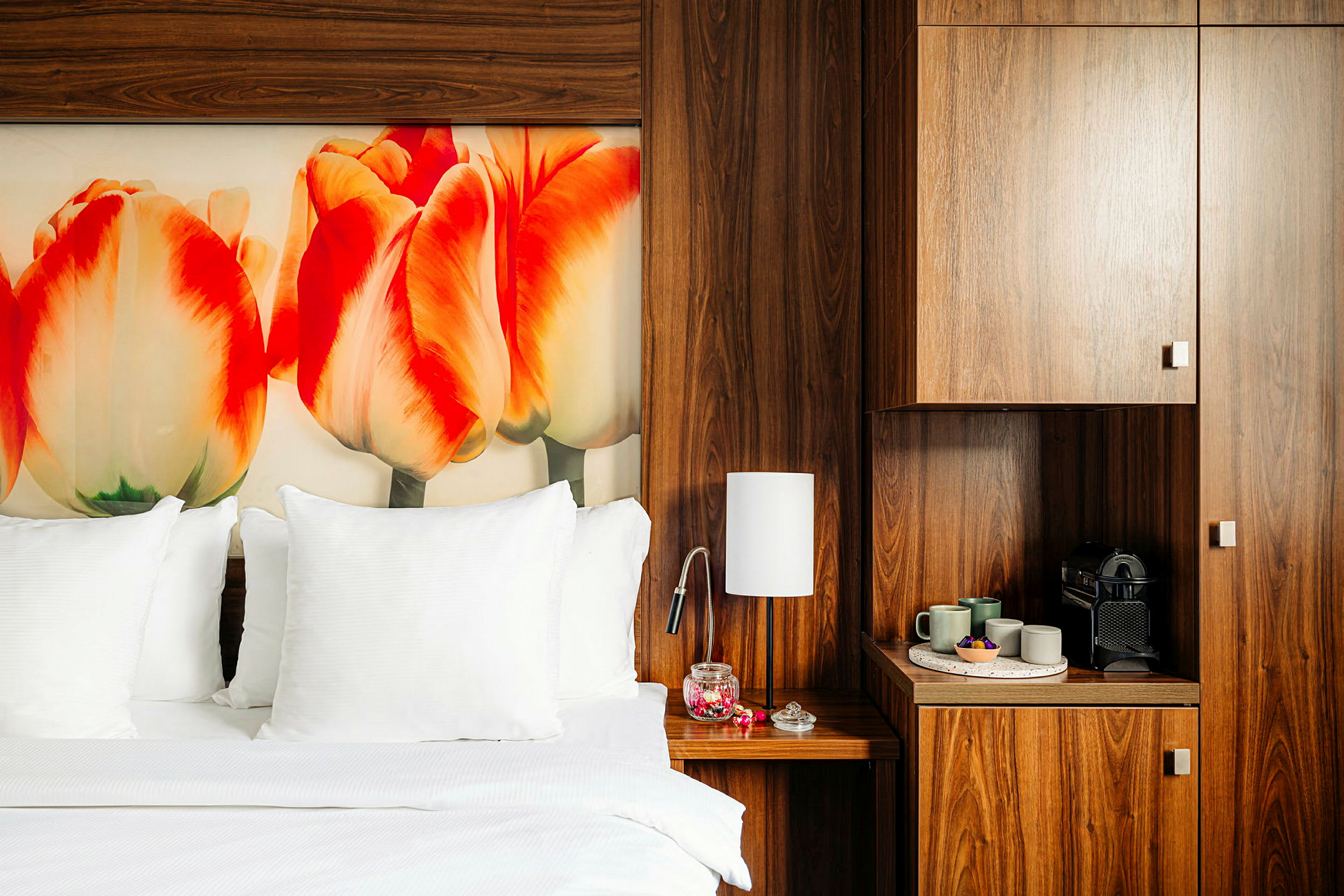 Hotellihuone, jossa on suuri sänky valkoisilla lakanoilla ja tyynyillä. Seinällä on värikäs kuva tulppaaneista. Huoneessa on tummia puuhuonekaluja, kuten yöpöytä lampulla ja kahvialue. (Tekoälyn luoma teksti)