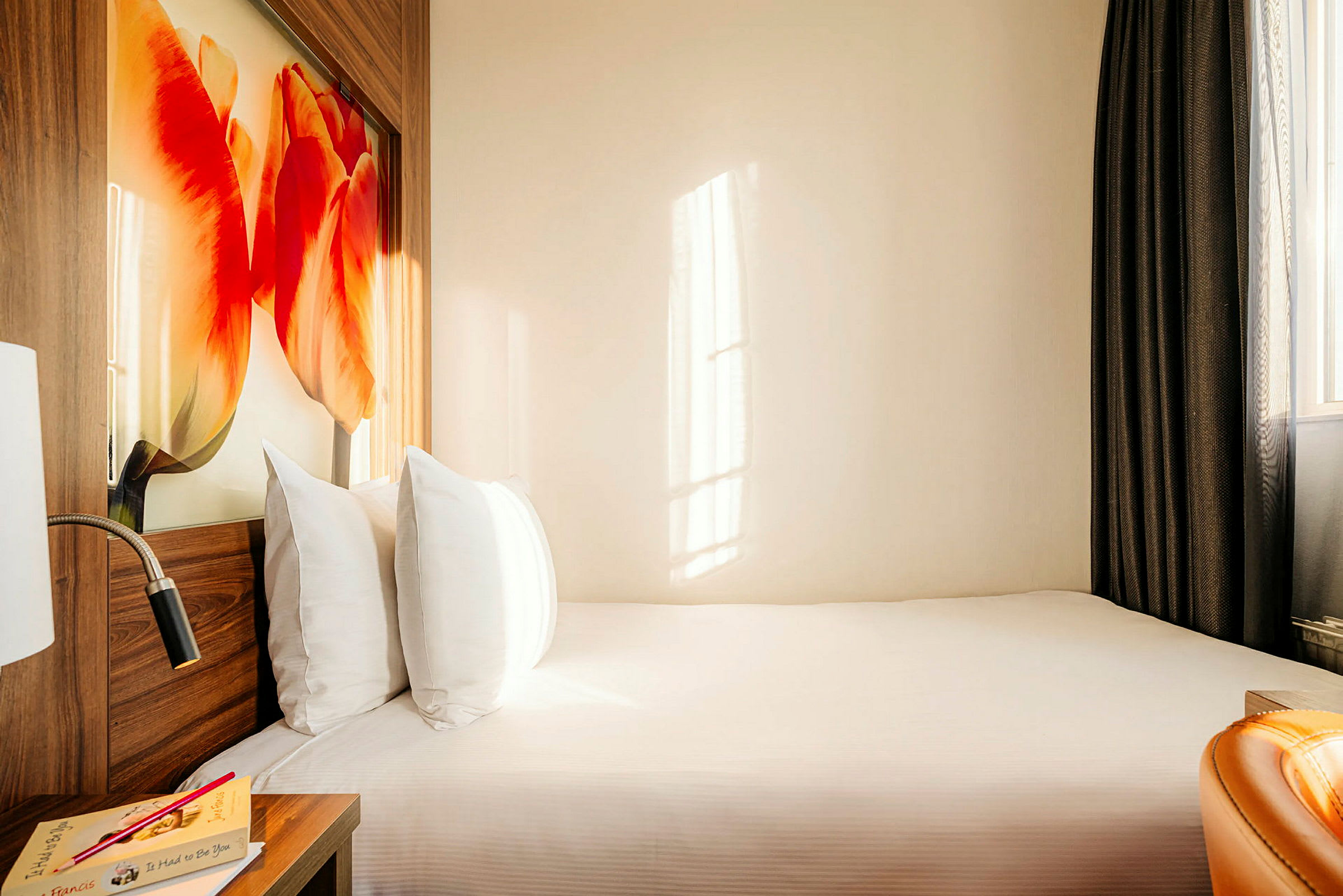 Hotellihuone, jossa sänky, peitto ja tyynyt, yöpöytä kirjan kanssa ja seinässä tulppaanikuvio. (Tekoälyn luoma teksti)