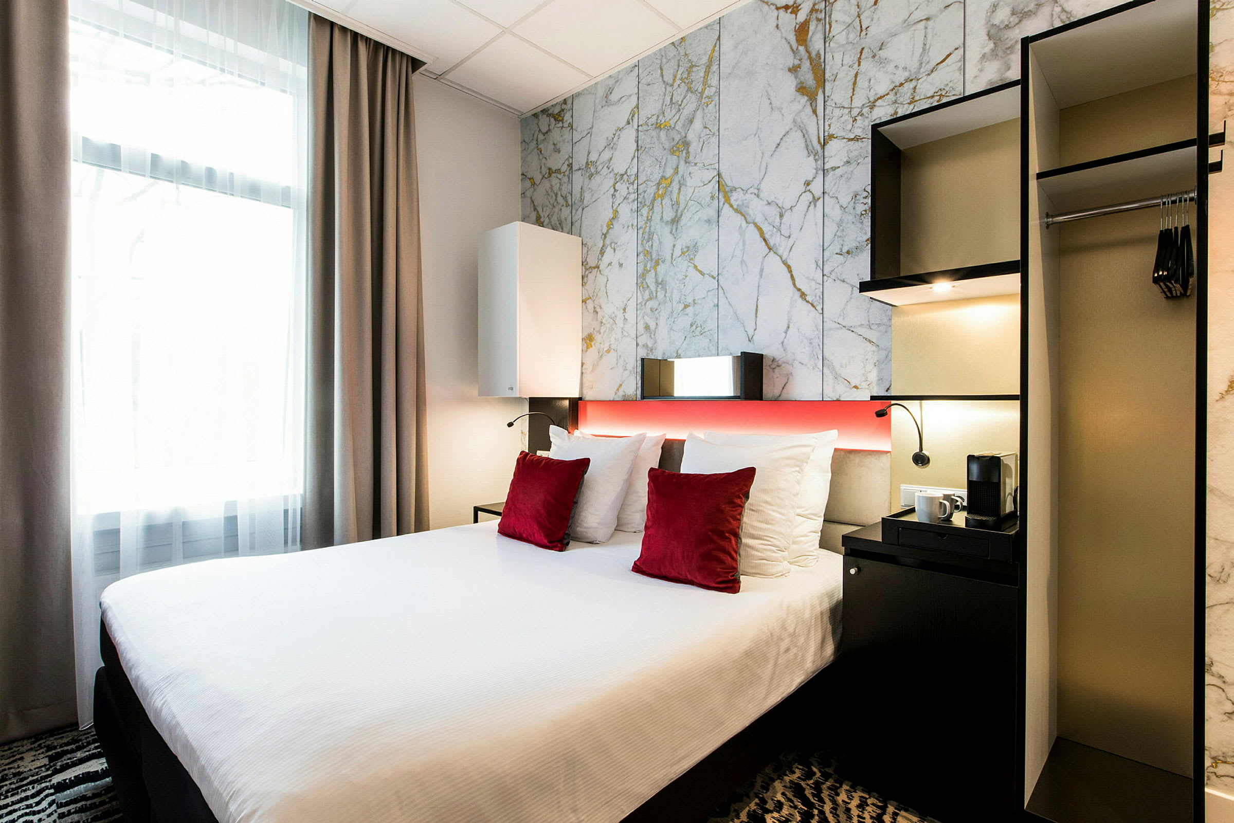 Hotellihuone, jossa on sänky, valkoiset vuodevaatteet ja punaiset koristeelliset tyynyt, vieressä kaappi ja pieni pöytä kahvinkeittimen kanssa. (Tekoälyn luoma teksti)
