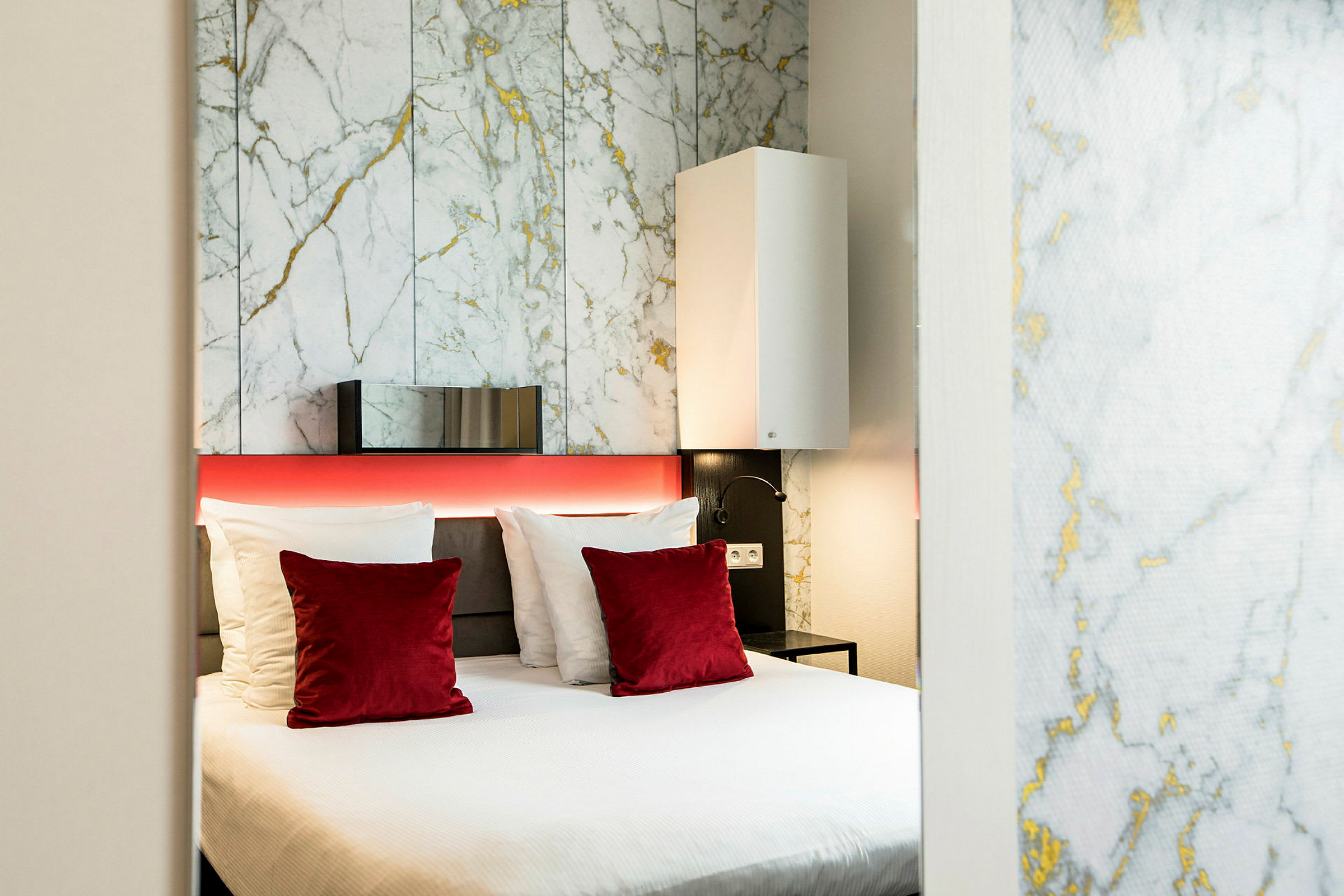 Moderni hotellihuone, jossa on parisänky, valkoisia ja punaisia tyynyjä sekä taustaseinässä marmorikuvio. (Tekoälyn luoma teksti)