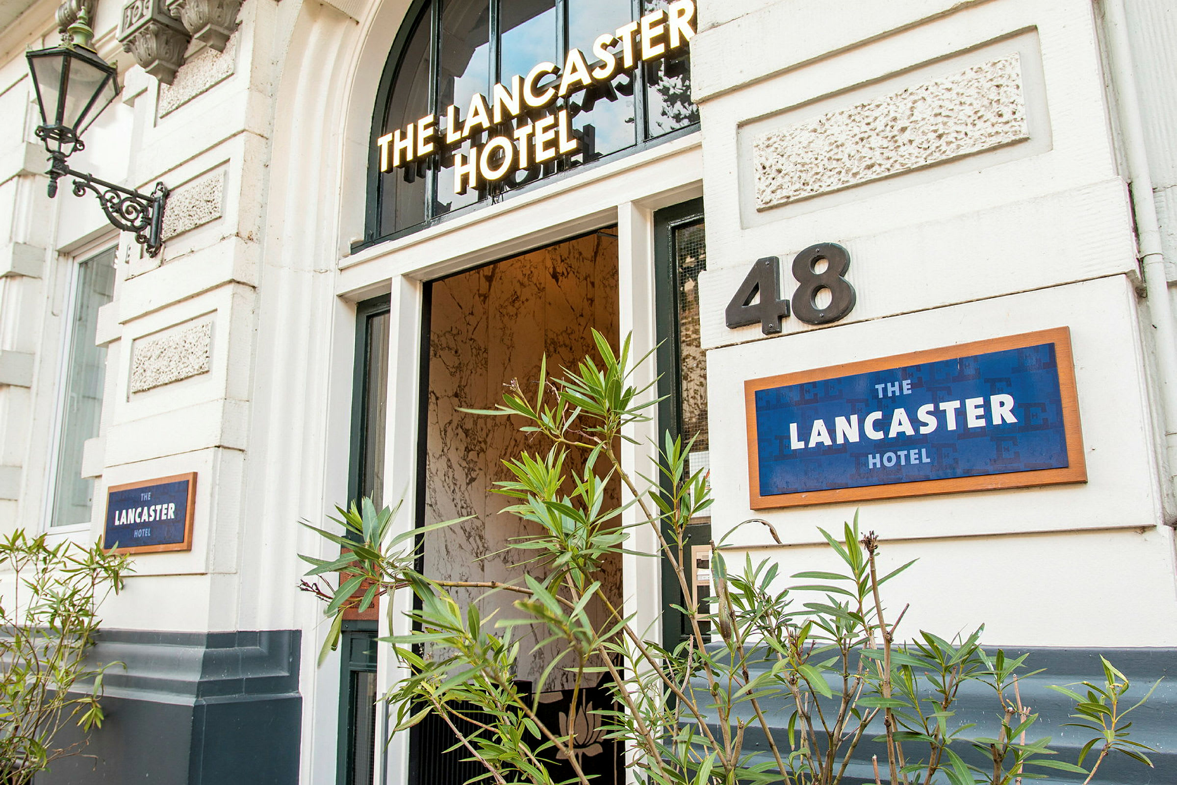 Hotellin sisäänkäynti, nimeltään 'The Lancaster Hotel', jota ympäröivät vihreät kasvit. (Tekoälyn luoma teksti)