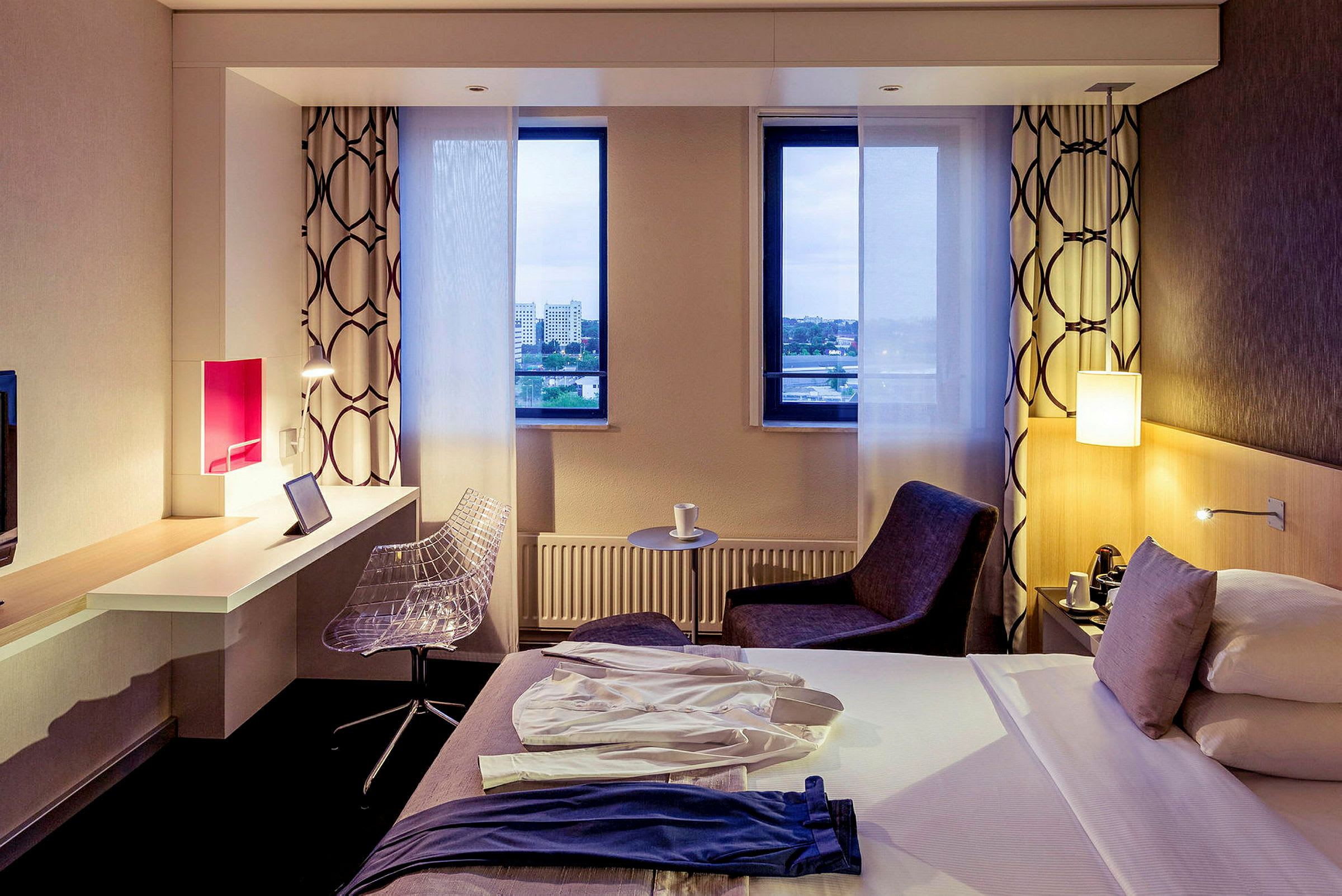 Hotellihuone, jossa on sänky, kirjoituspöytä ja tuoli sekä kaksi ikkunaa, joista on näkymä. (Tekoälyn luoma teksti)