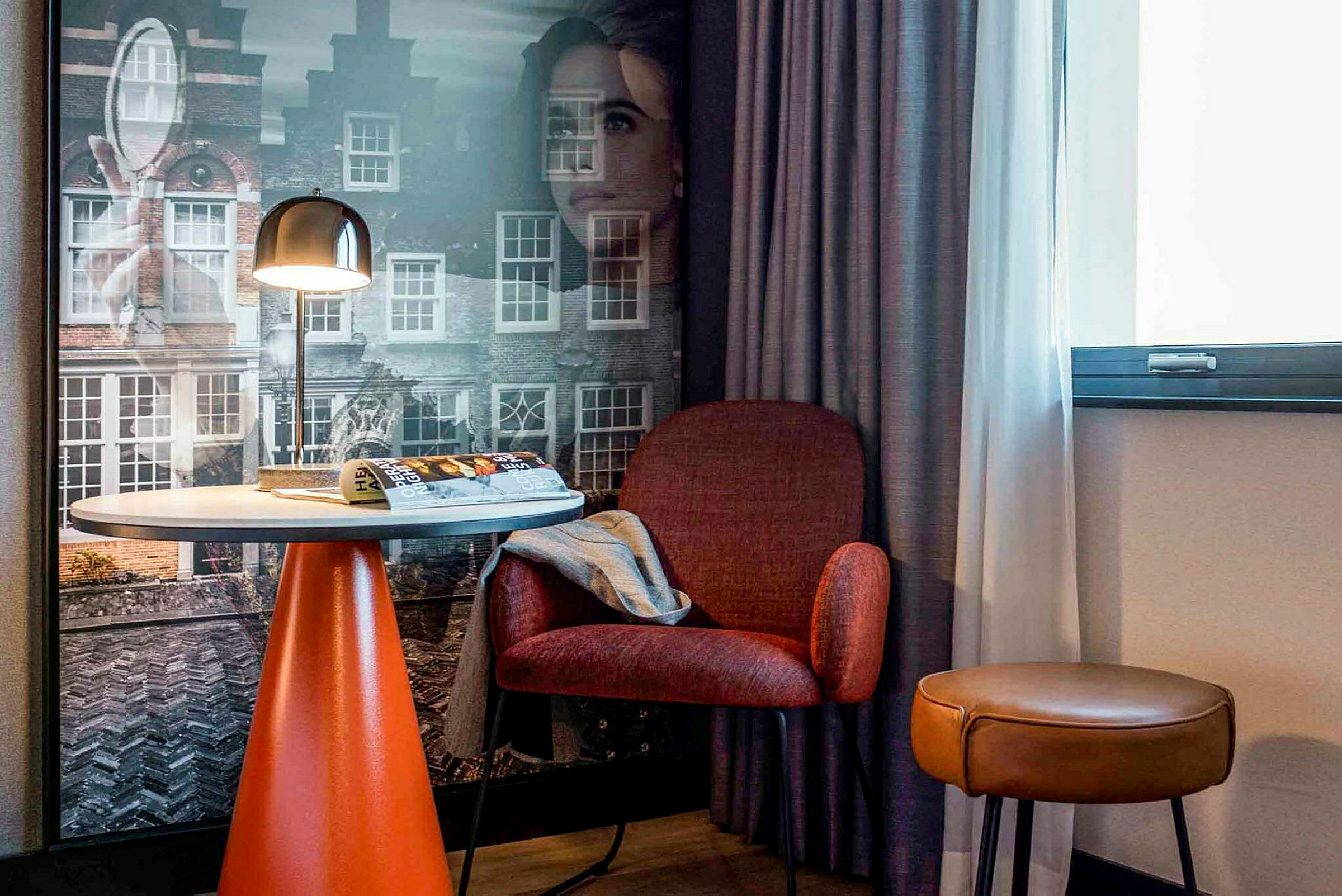 Hotellihuone, jossa pöytä, punainen tuoli ja ruskea jakkara ikkunan edessä ja seinä kaupunkimotiiveilla. (Tekoälyn luoma teksti)