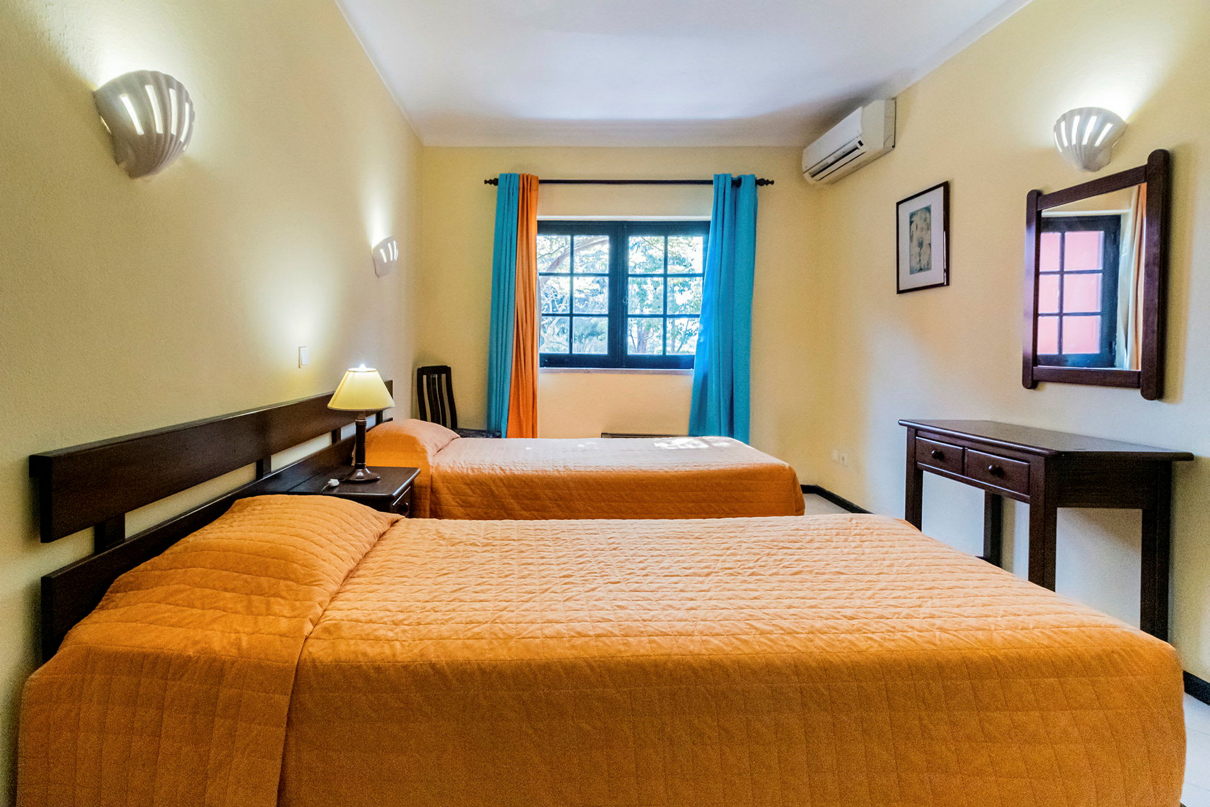Hotellihuone, jossa on kaksi erillistä sänkyä, seinäpeili, kirjoituspöytä ja ikkuna värikkäillä verhoilla. (Tekoälyn luoma teksti)