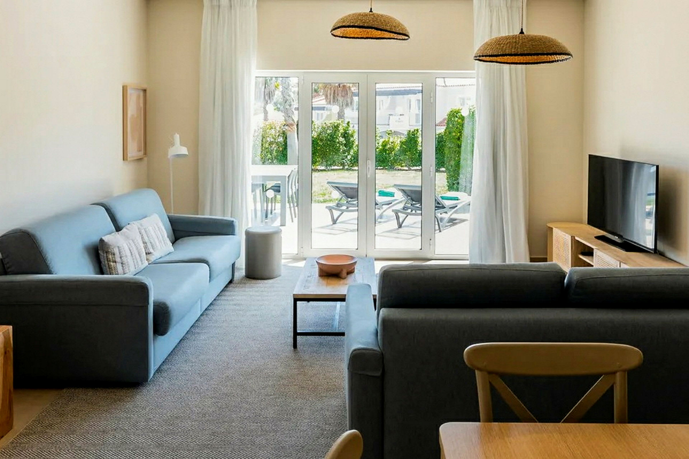 Olohuone, jossa on kaksi sohvaa, sohvapöytä, televisio ja lasiovi, joka avautuu terassille, jossa on aurinkotuoleja. (Tekoälyn luoma teksti)
