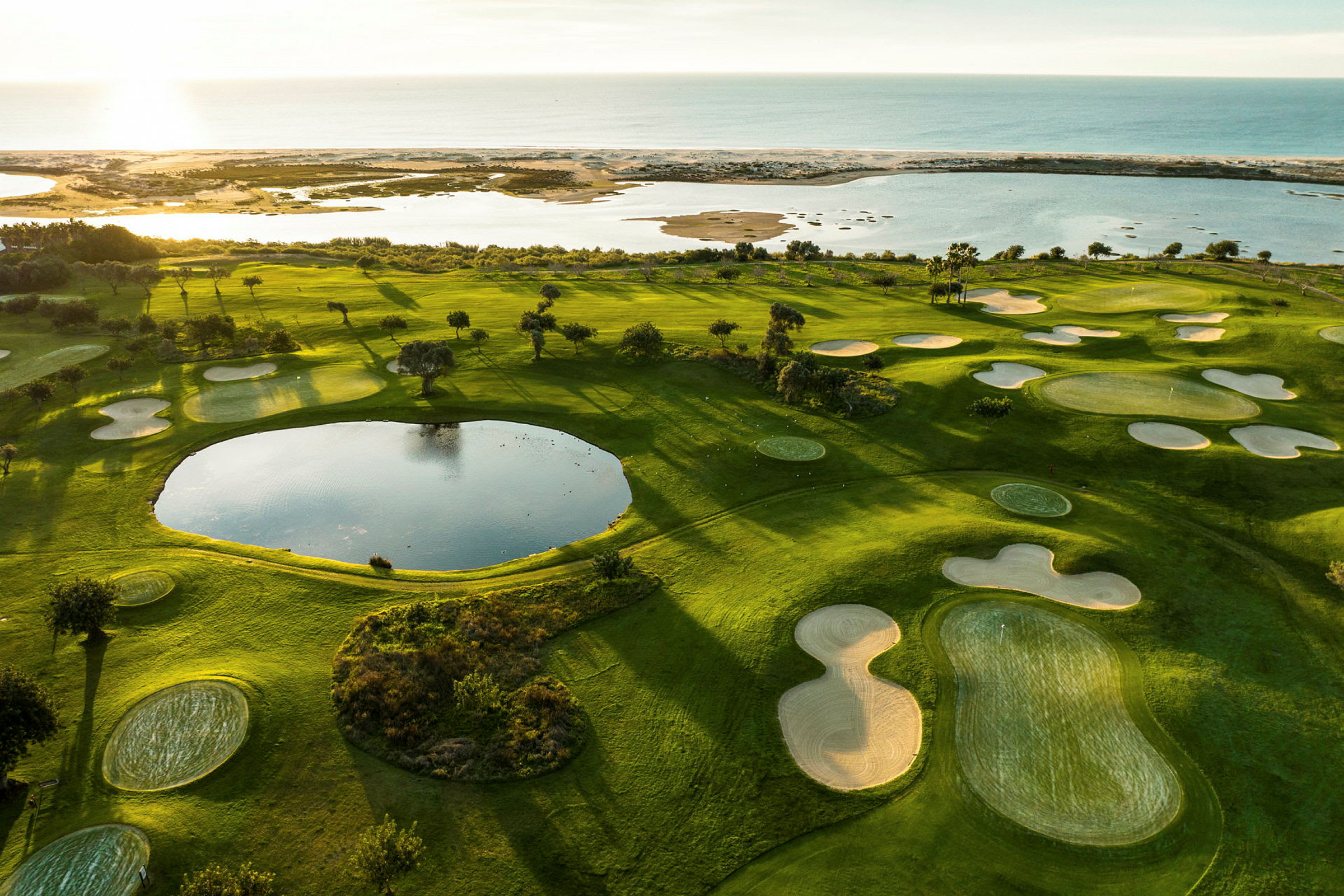 Näkymä vihreälle golfkentälle, jossa hiekkasyvennyksiä ja lampi, lähellä rannikkoa ja merta. (Tekoälyn luoma teksti)