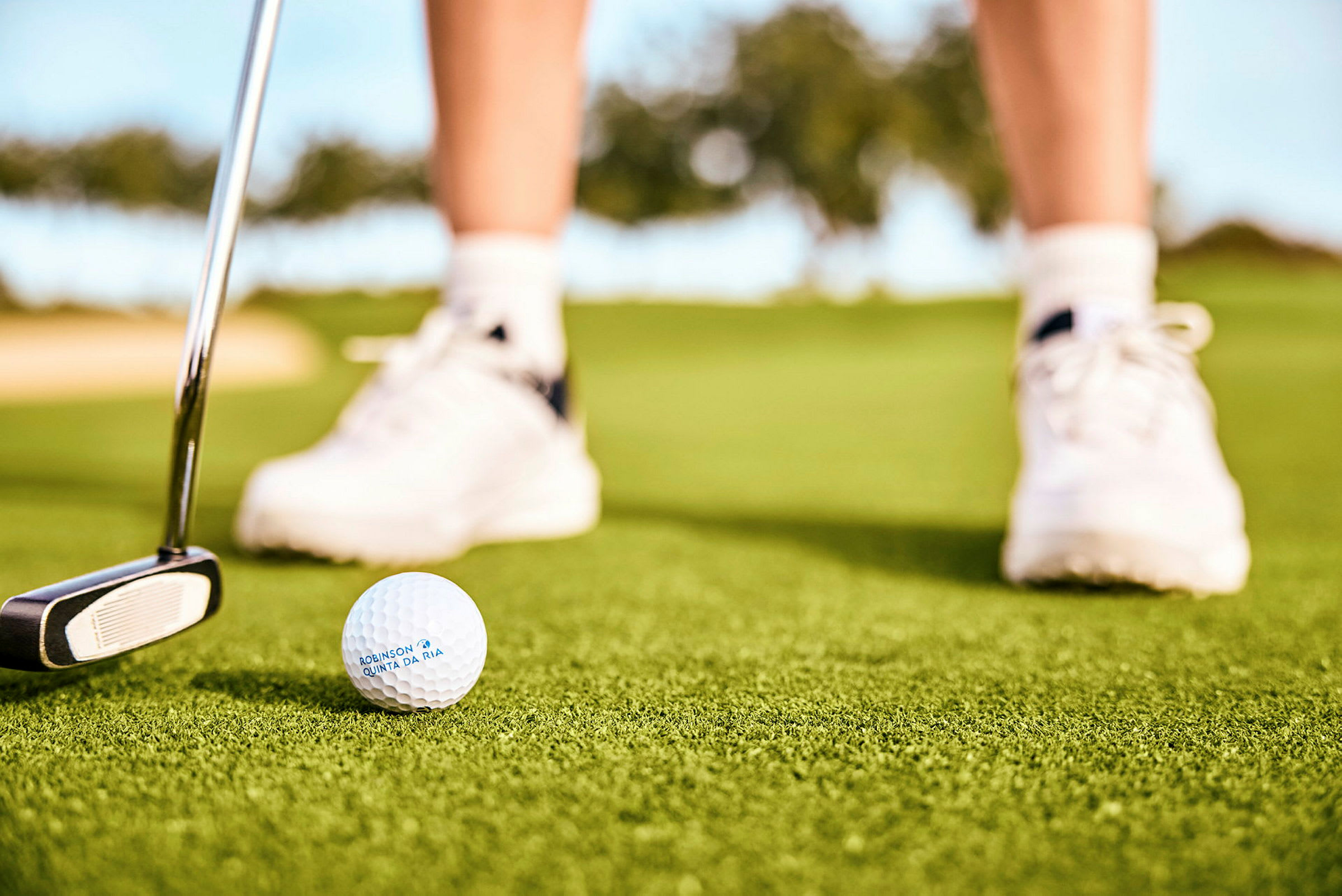 Golfpallo ja putteri vihreällä nurmikolla, taustalla henkilön jalat, joilla on valkoiset urheilukengät. (Tekoälyn luoma teksti)