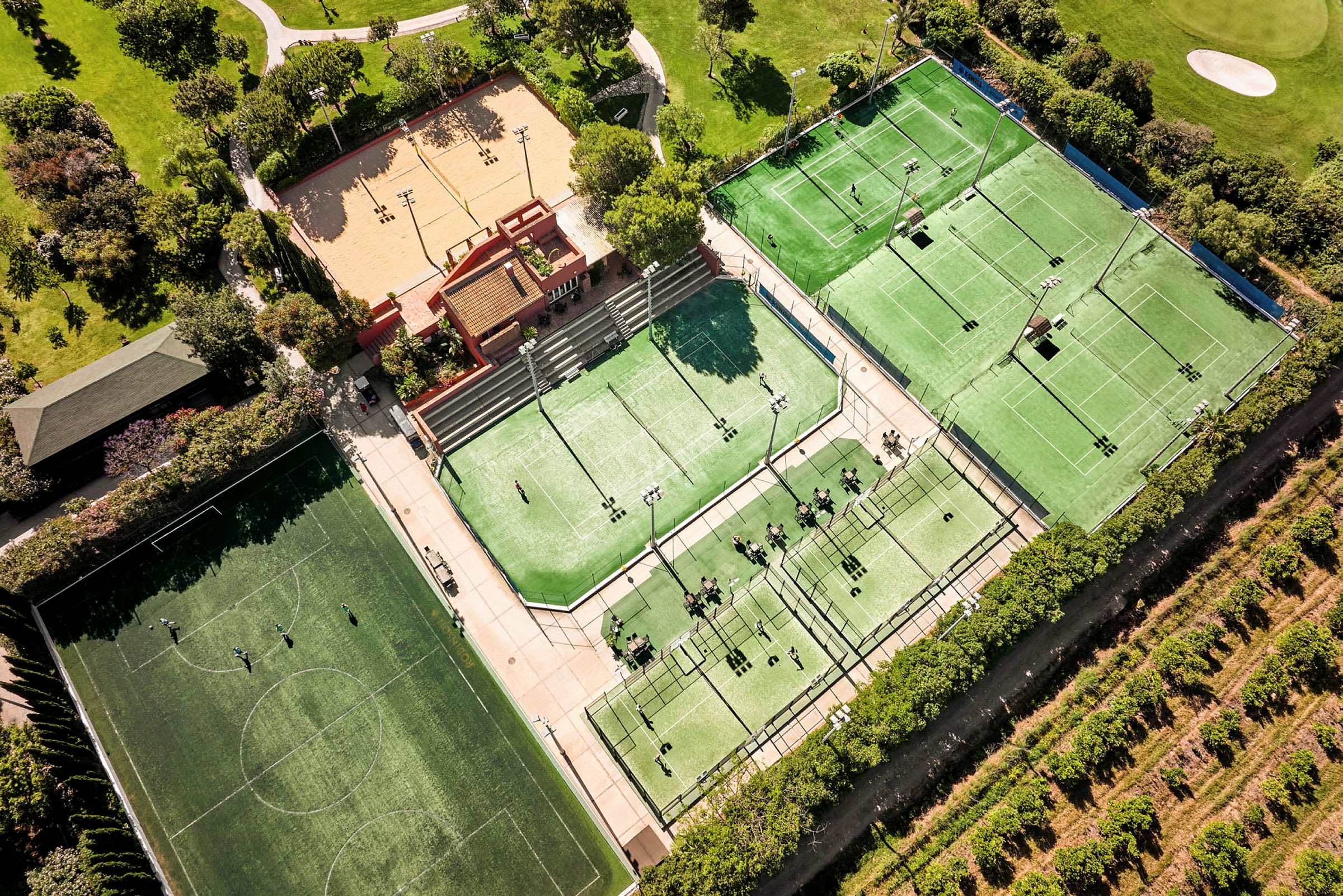 Ilmakuvassa urheilukeskus, jossa on tenniskenttiä, padelkenttiä, jalkapallokenttä ja hiekkakenttä, ympäröitynä vihreydellä. (Tekoälyn luoma teksti)