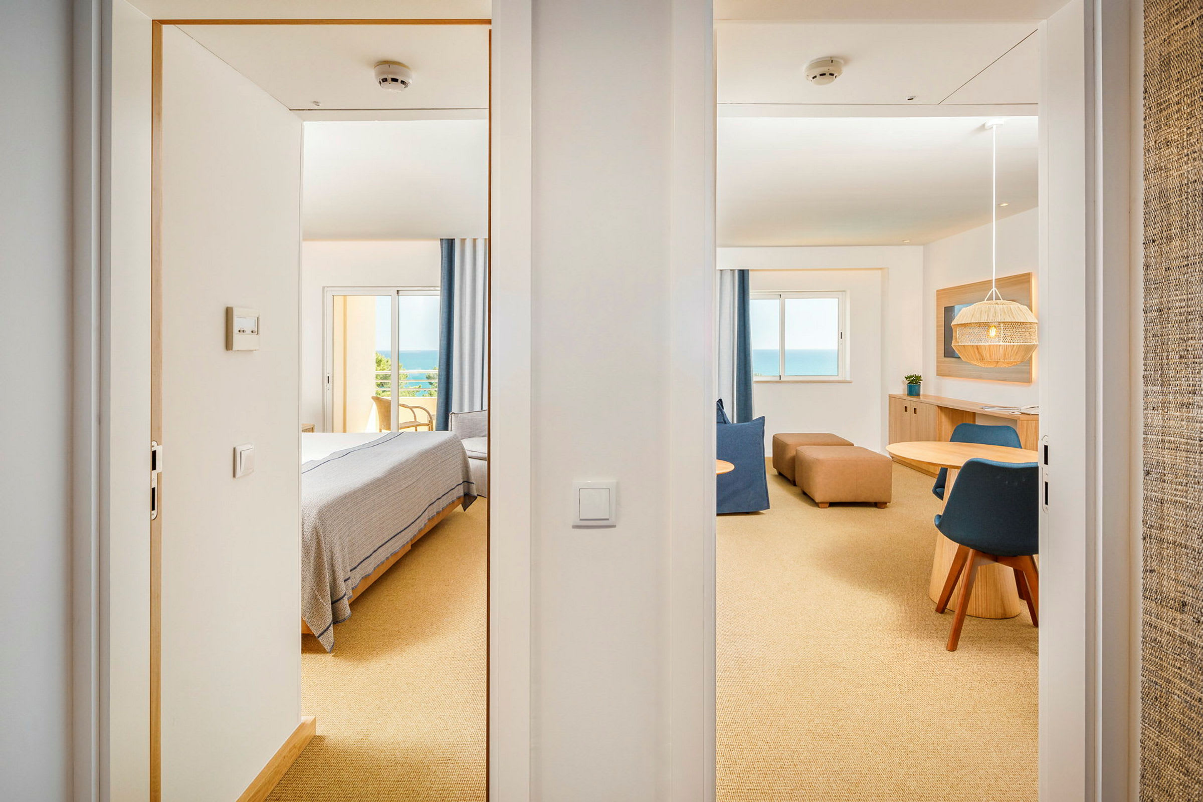 Näköala kahden oviaukon läpi valoiseen hotellihuoneeseen, jossa on makuualue ja oleskelutila sekä suuri ikkuna merinäköalalla. (Tekoälyn luoma teksti)