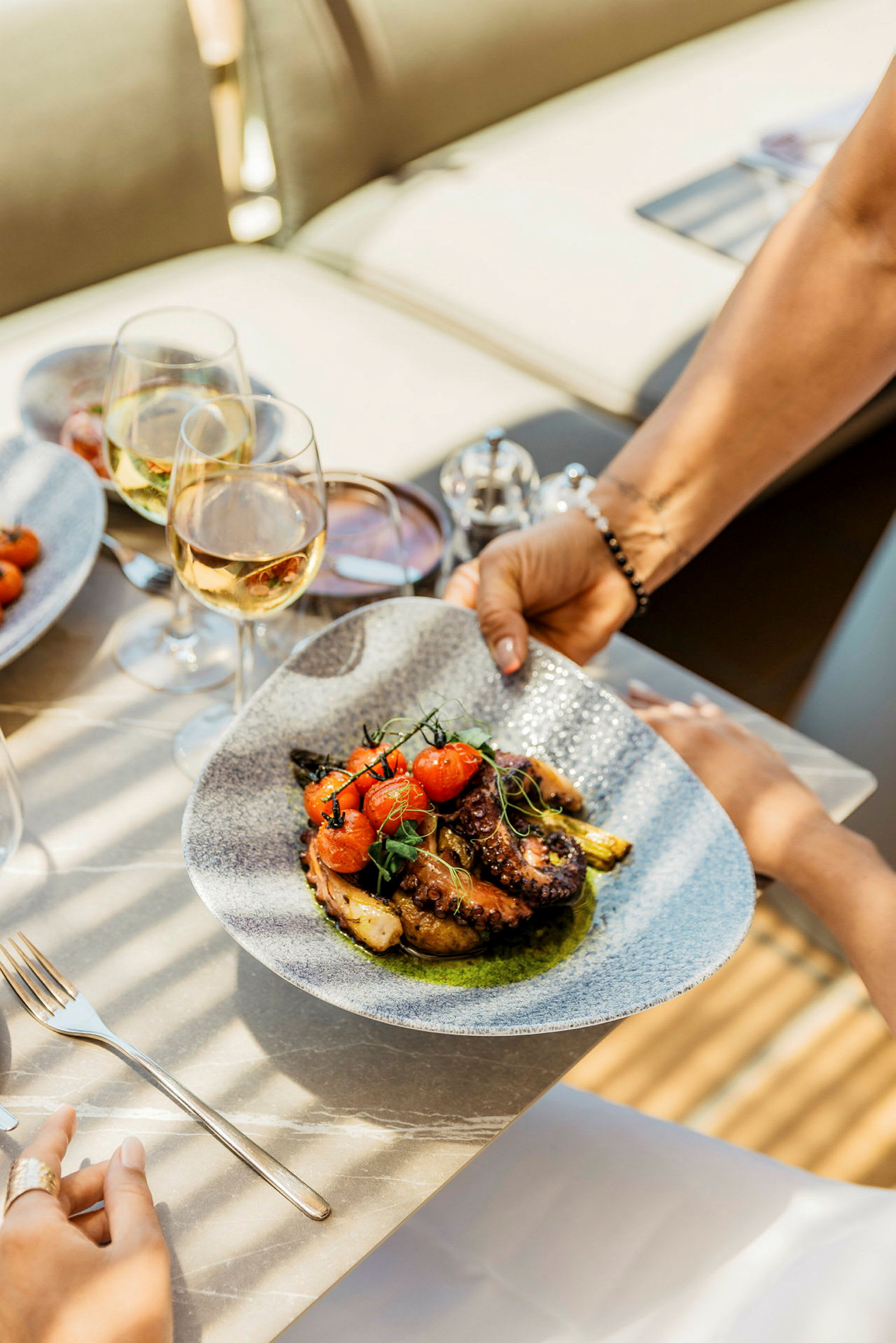 Kuvassa näkyy lautanen, jolla on herkullinen ruoka-annos sisältäen grillattua mustekalaa, tomaatteja ja vihanneksia, tarjolla pöydässä viinilasien kanssa. (Tekoälyn luoma teksti)