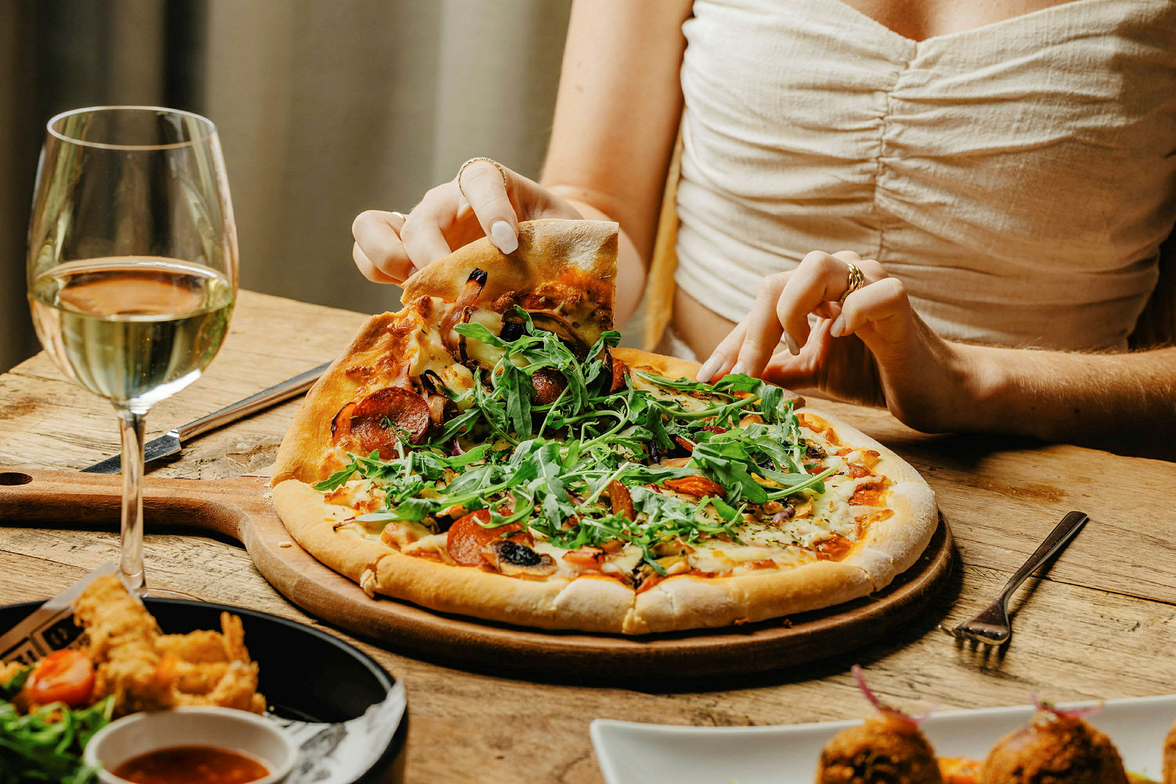 Lähikuva henkilöstä, joka ottaa pizzapalan lautaselta, vieressä lasi valkoviiniä. (Tekoälyn luoma teksti)