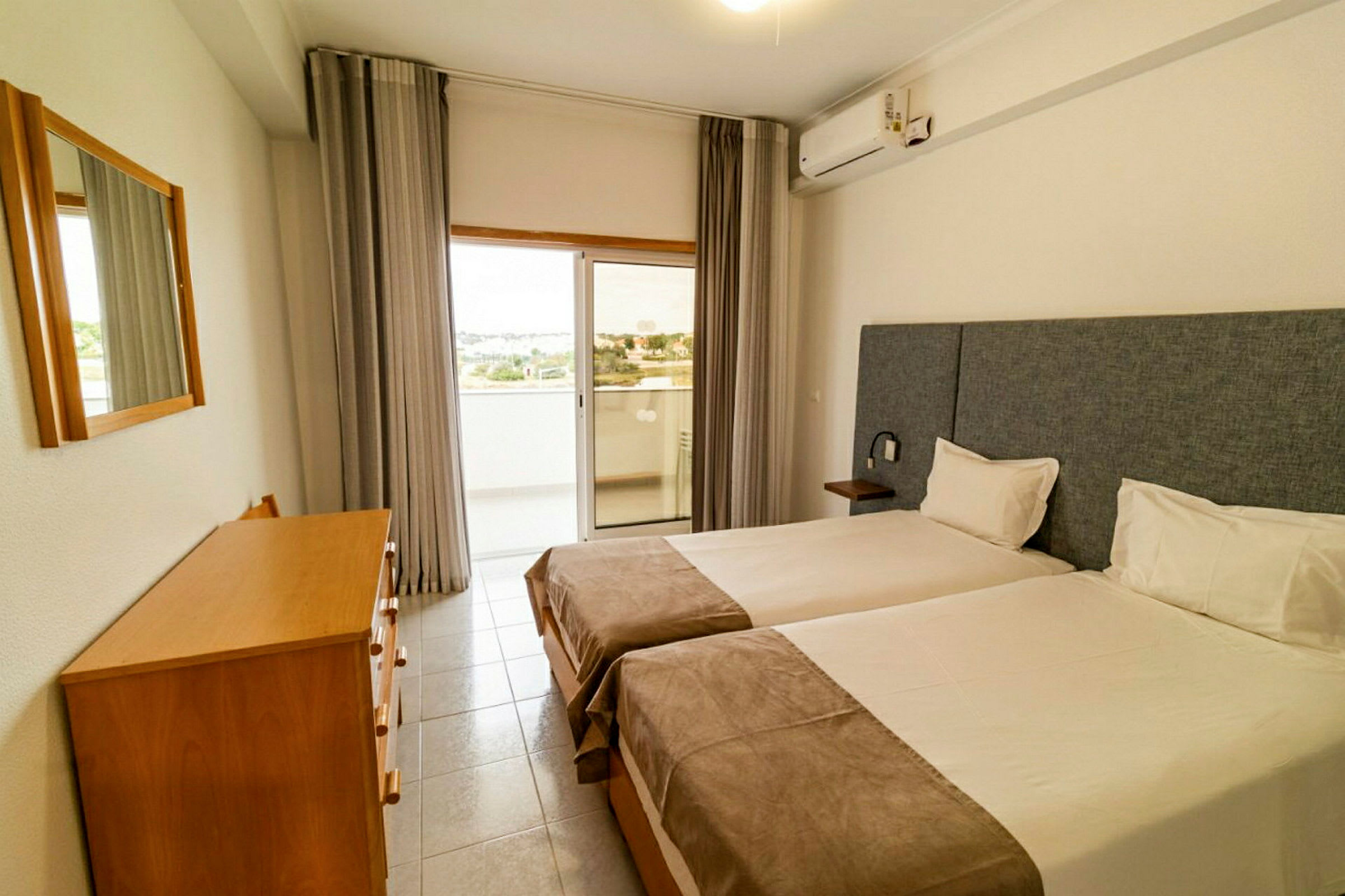 Hotellihuone, jossa on kaksi yhden hengen vuodetta, lipasto ja peili sekä lasiovi, joka johtaa parvekkeelle. (Tekoälyn luoma teksti)