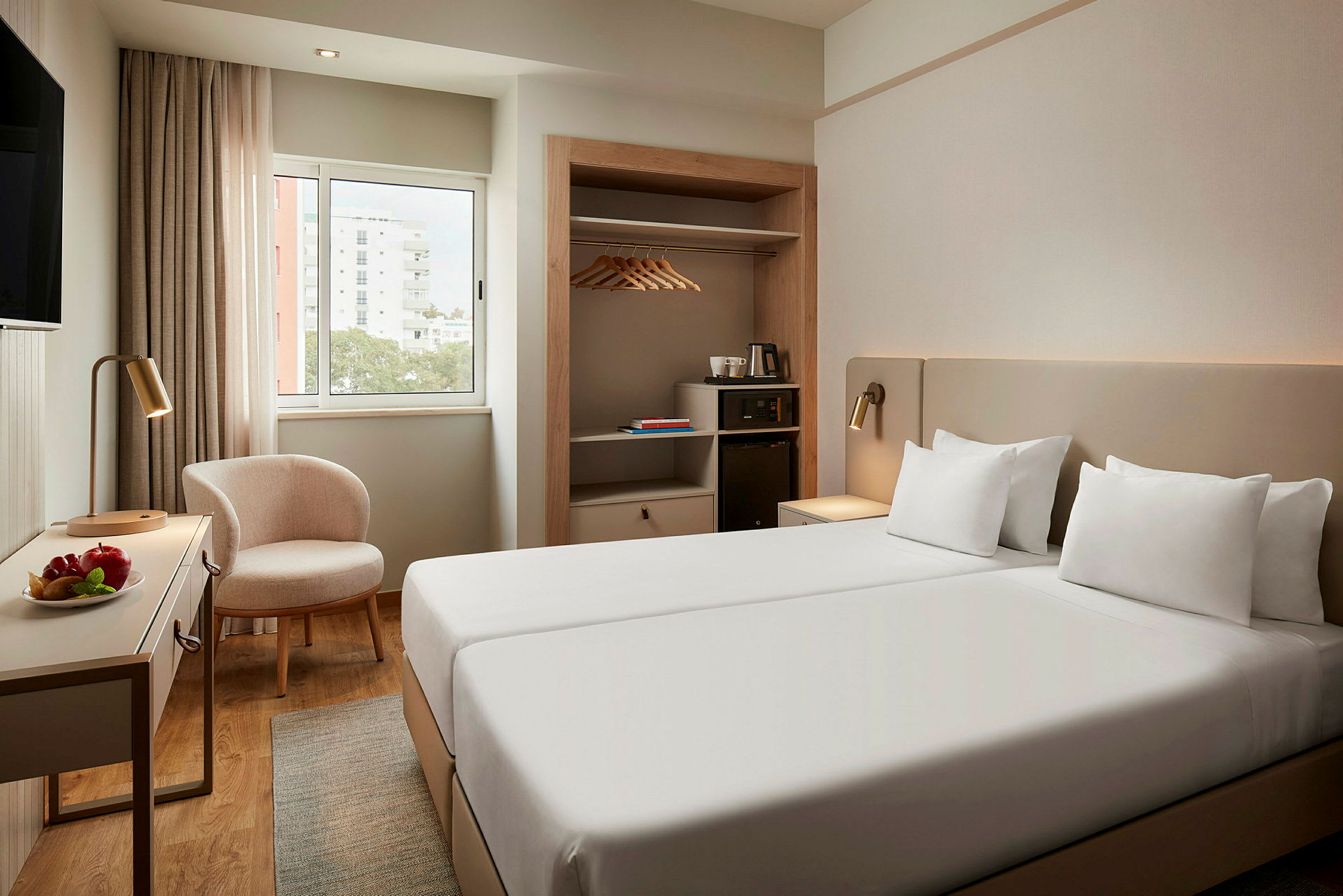 Hotellihuone, jossa on kaksi yksittäissänkyä, ikkuna, tuoli, pöytä hedelmäkulholla ja kaappi vaateripustimilla. (Tekoälyn luoma teksti)