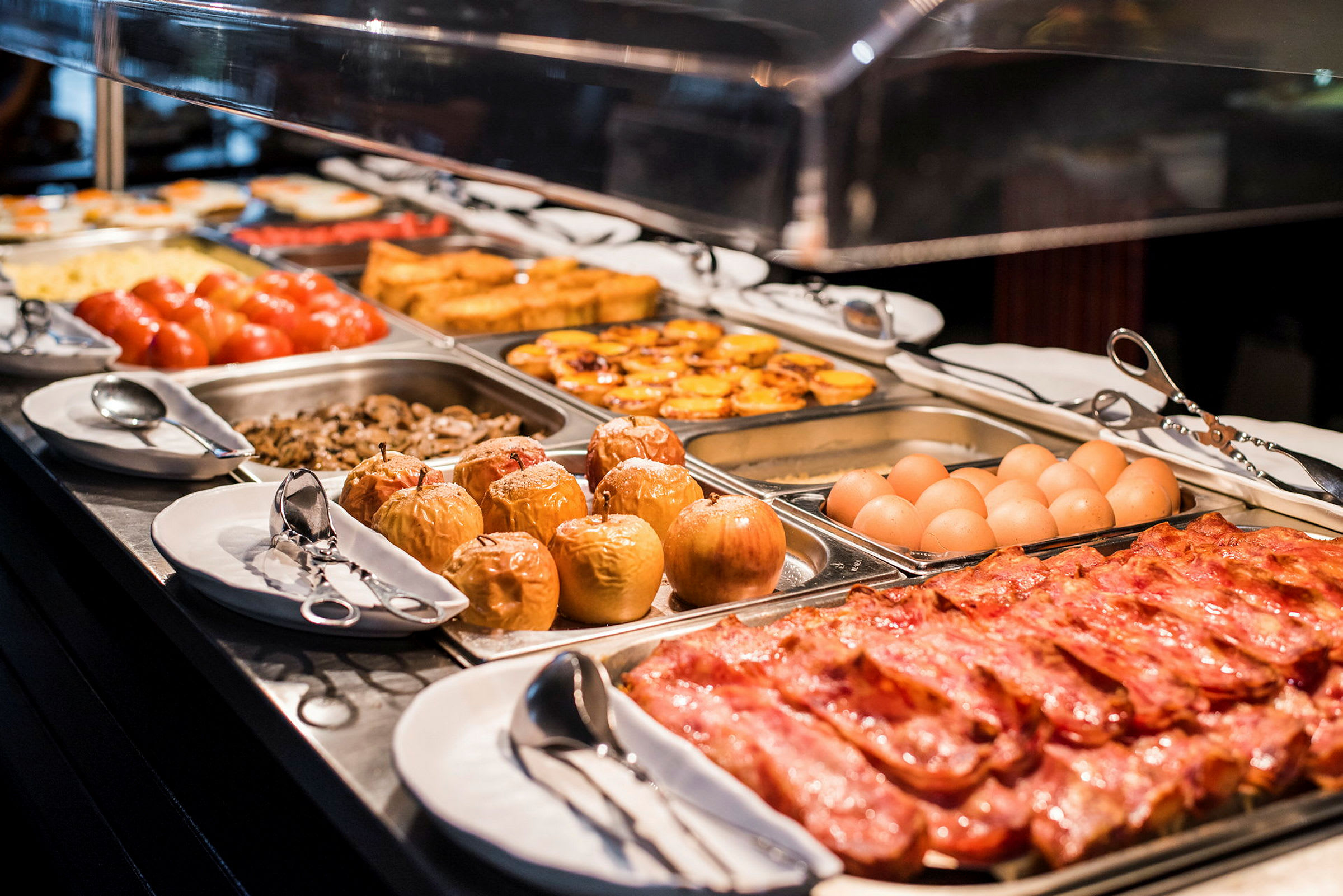 Buffet-pöytä, jossa on erilaisia ruokia, kuten pekonia, kananmunia, uunissa paistettuja omenoita, tomaatteja ja laatikoita. (Tekoälyn luoma teksti)