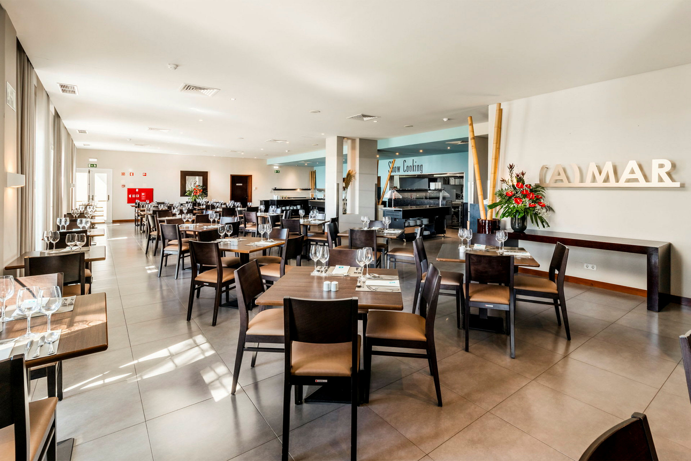 Valoisa ja moderni ravintola, jossa on siististi järjestetyt pöydät ja tuolit, raikkaita kukkia ja suuret ikkunat. (Tekoälyn luoma teksti)