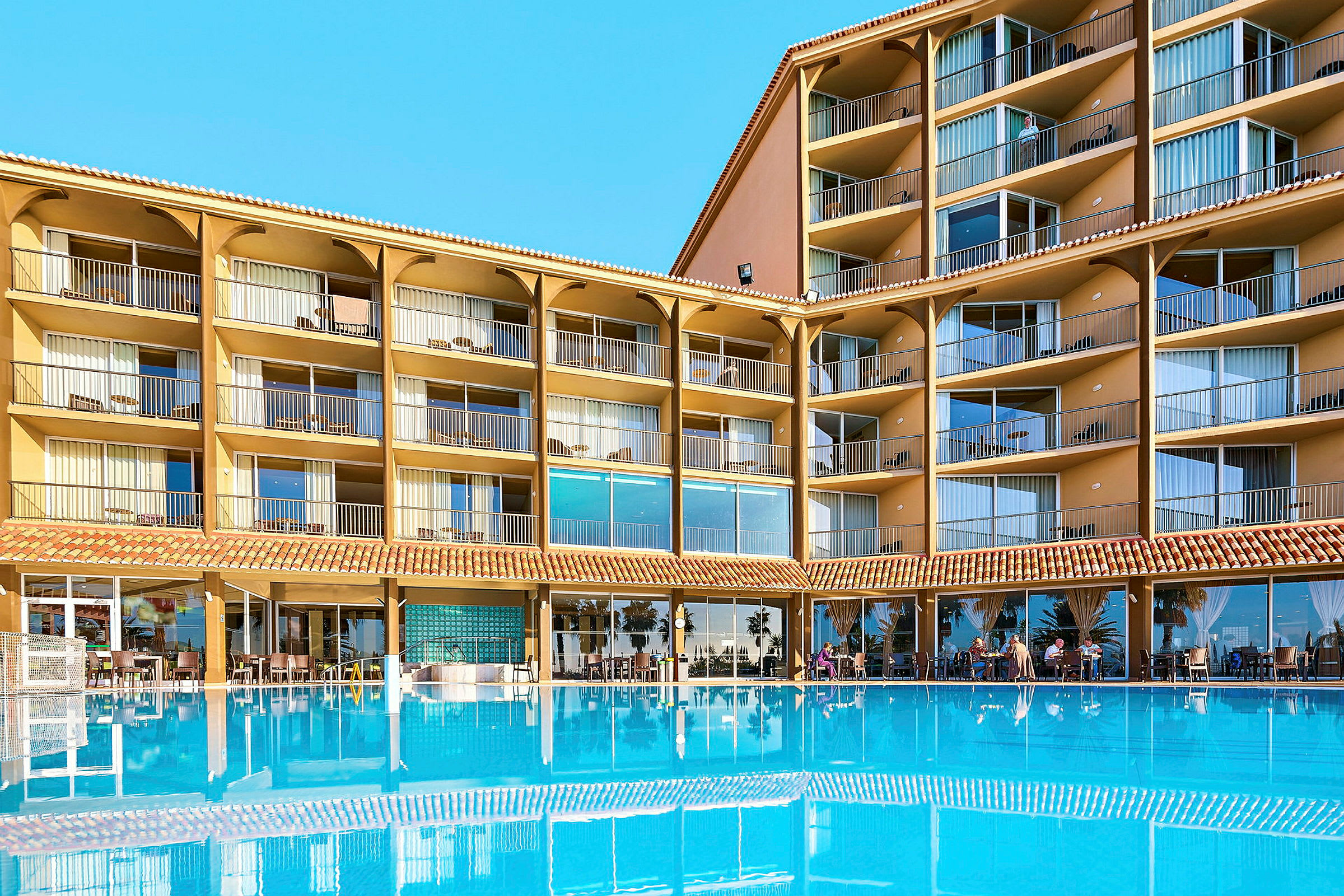 Hotellirakennus, jossa on useita kerroksia ja parvekkeita, suuren sinisen uima-altaan edessä. (Tekoälyn luoma teksti)