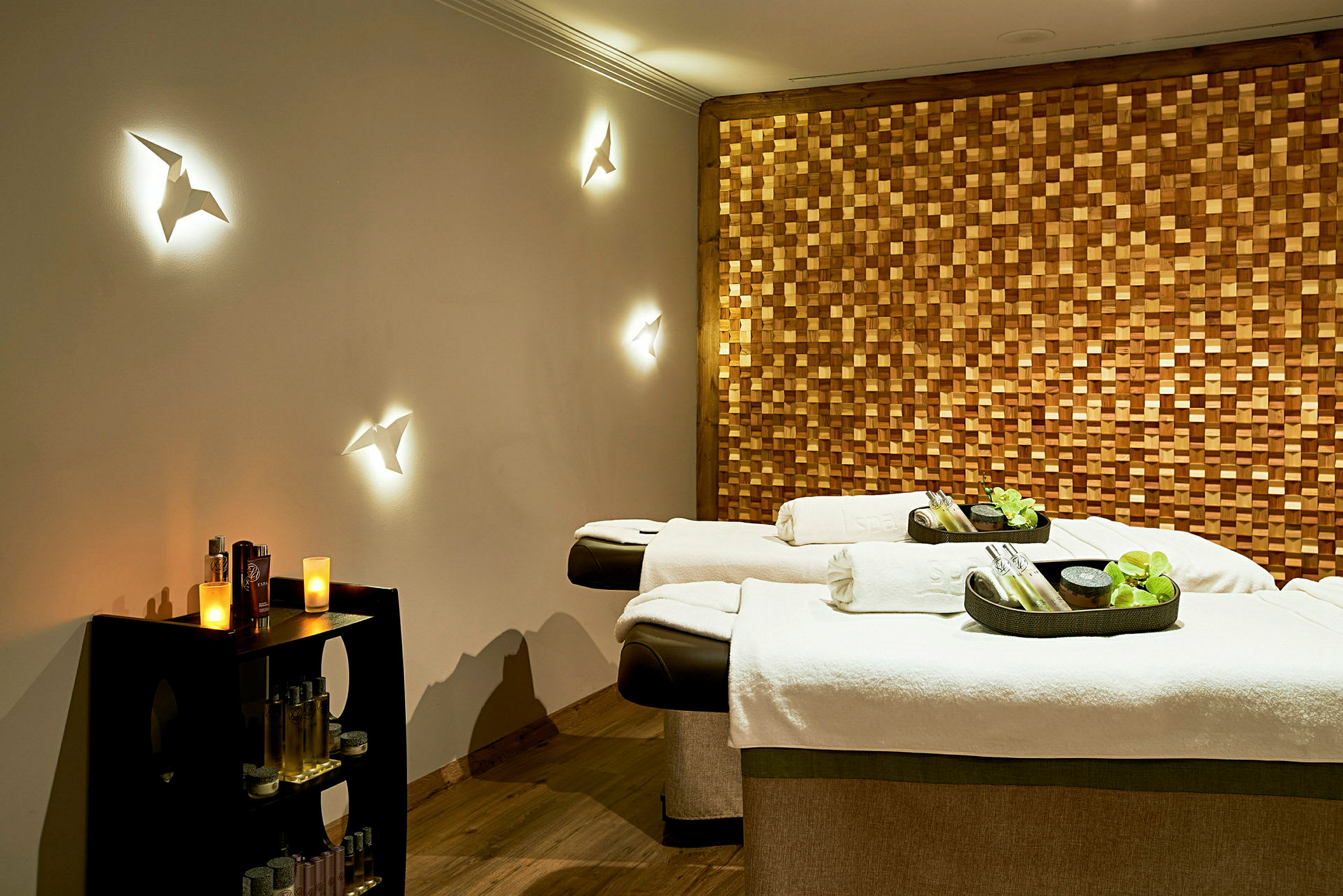 Spa-huone, jossa on kaksi hierontapöytää, koristeelliset seinävalaisimet ja puinen seinä. (Tekoälyn luoma teksti)