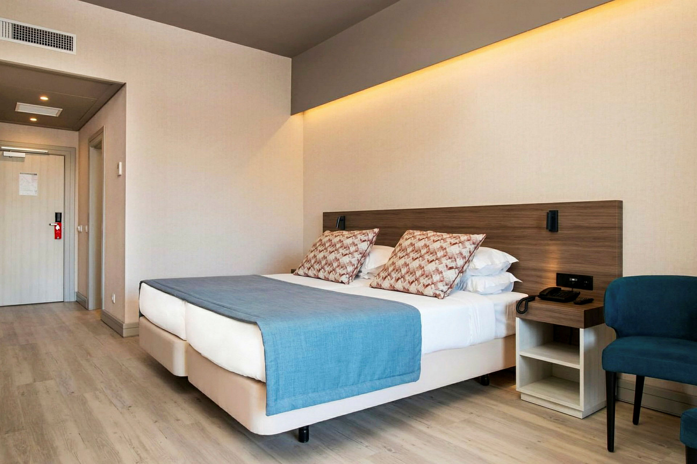 Hotellihuone, jossa on parivuode, sininen huopa ja tyynyjä, vieressä tuoli ja yöpöytä. (Tekoälyn luoma teksti)