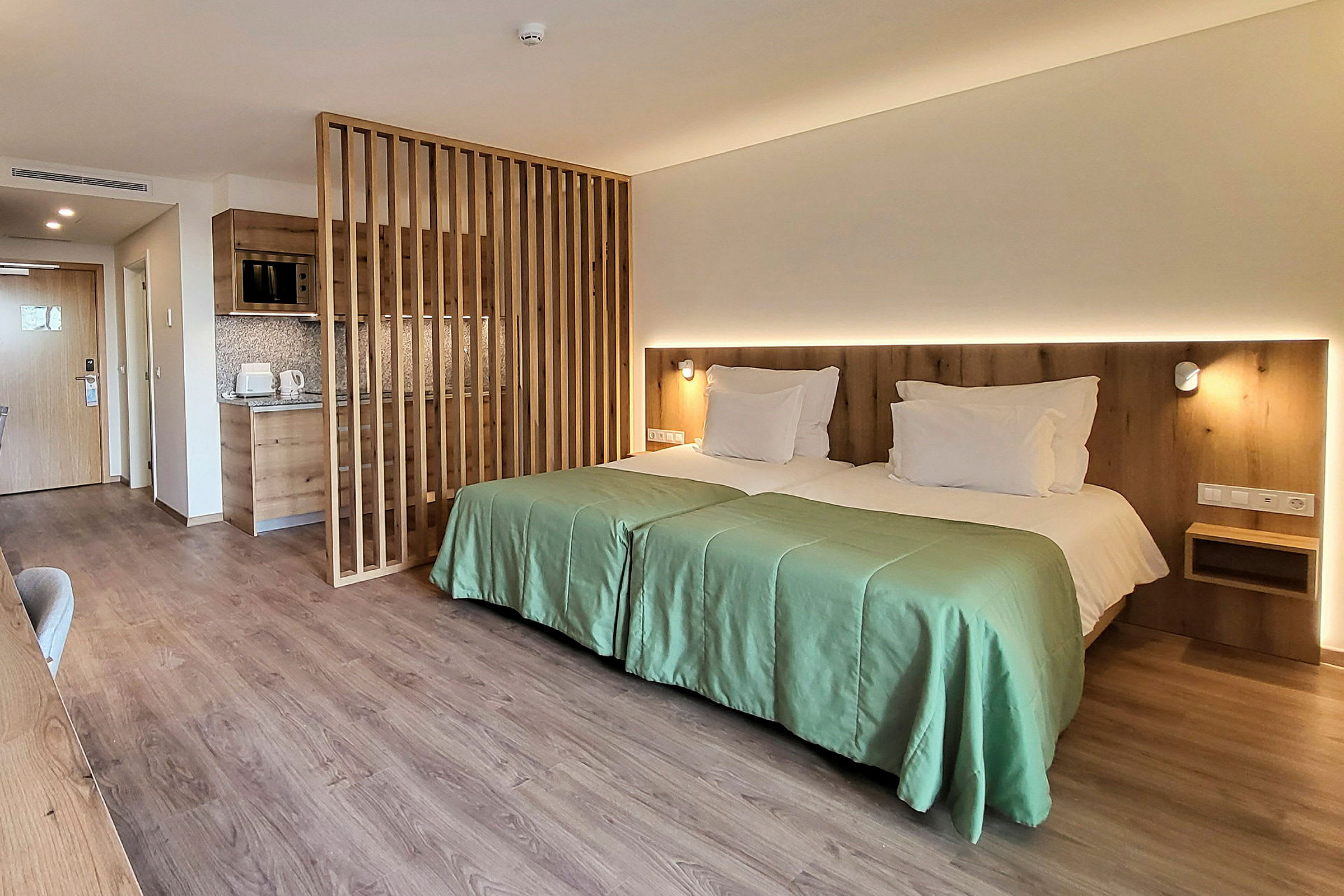 Moderni hotellihuone, jossa kaksi vierekkäistä erillissänkyä, puupaneeliseinä ja pieni keittonurkkaus. (Tekoälyn luoma teksti)