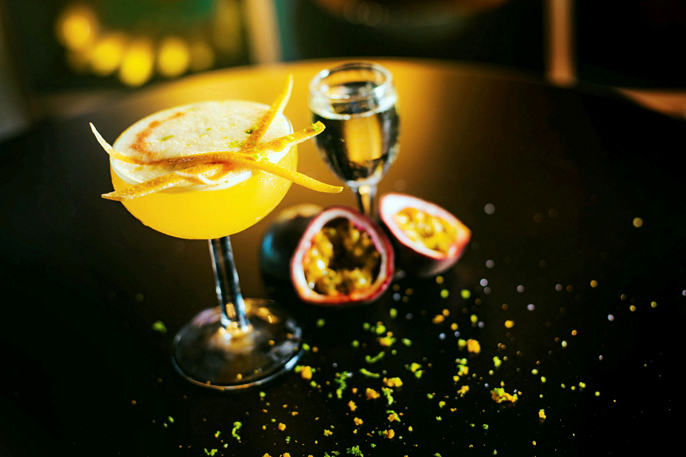 Keltainen cocktail koristeineen pöydällä, lasin ja halkaistun passionhedelmän vieressä. (Tekoälyn luoma teksti)
