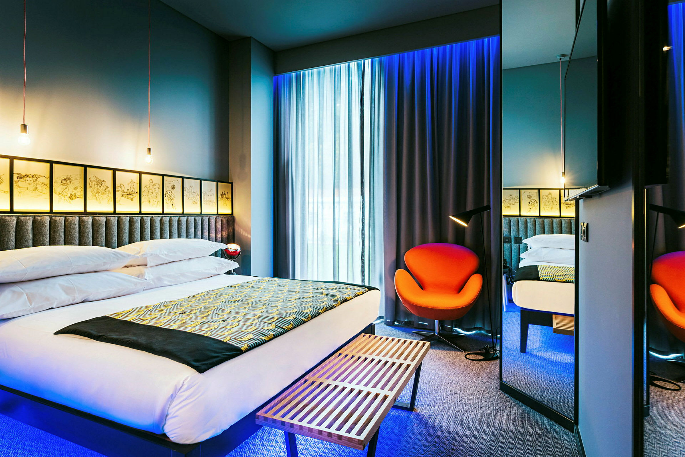 Moderni hotellihuone, jossa suuri sänky, oranssi tuoli ja pehmeästi valaistut verhot. (Tekoälyn luoma teksti)