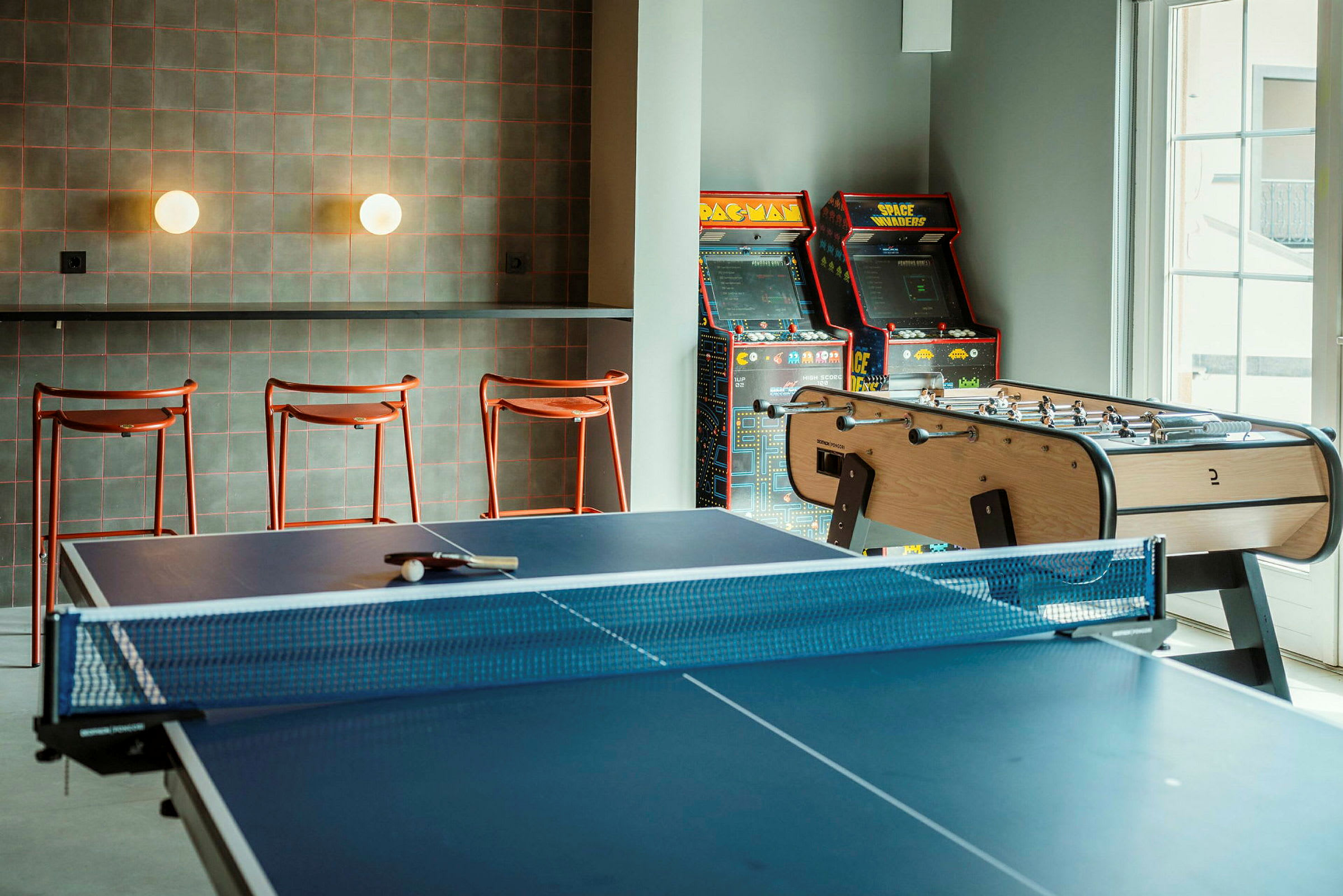 Huone, jossa on pöytätennispöytä, pöytäjalkapallopeli ja arcade-pelejä seinän vieressä. (Tekoälyn luoma teksti)
