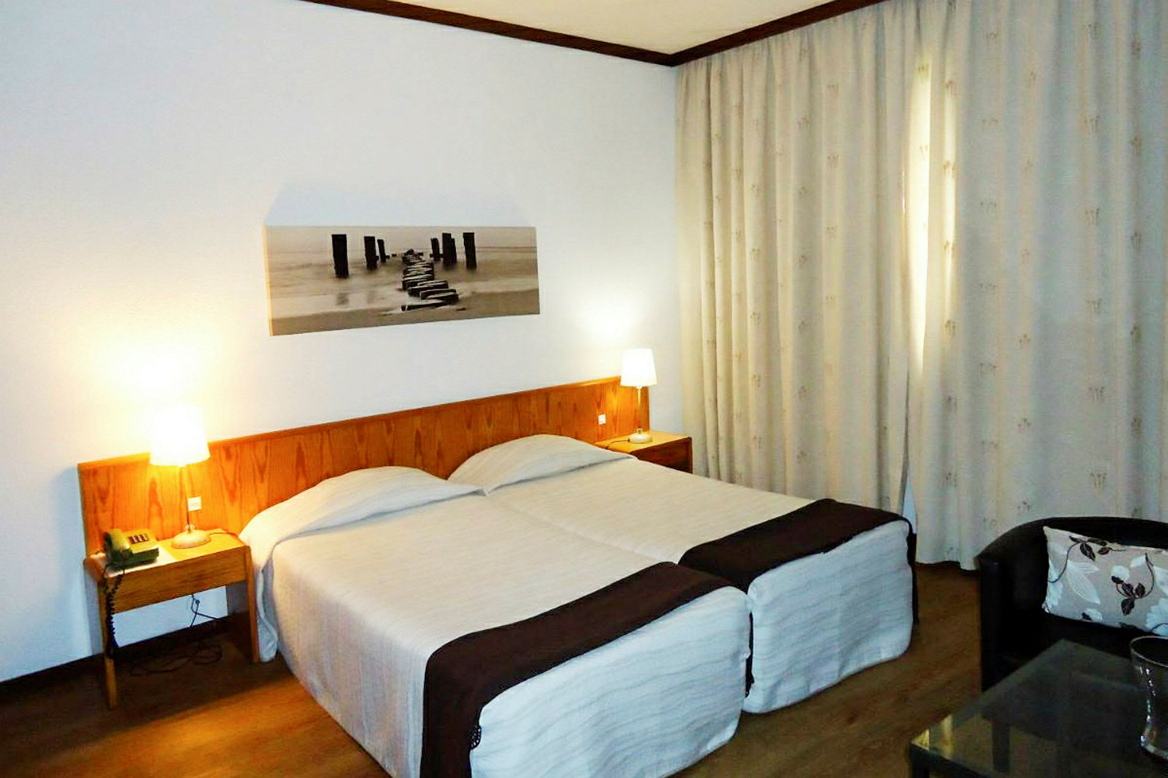 Hotellihuone, jossa on kaksi yhden hengen vuodetta, puinen sängynpääty ja yöpöydät lampuilla. Seinällä on taulu ja ikkunaa peittävät verhot. (Tekoälyn luoma teksti)