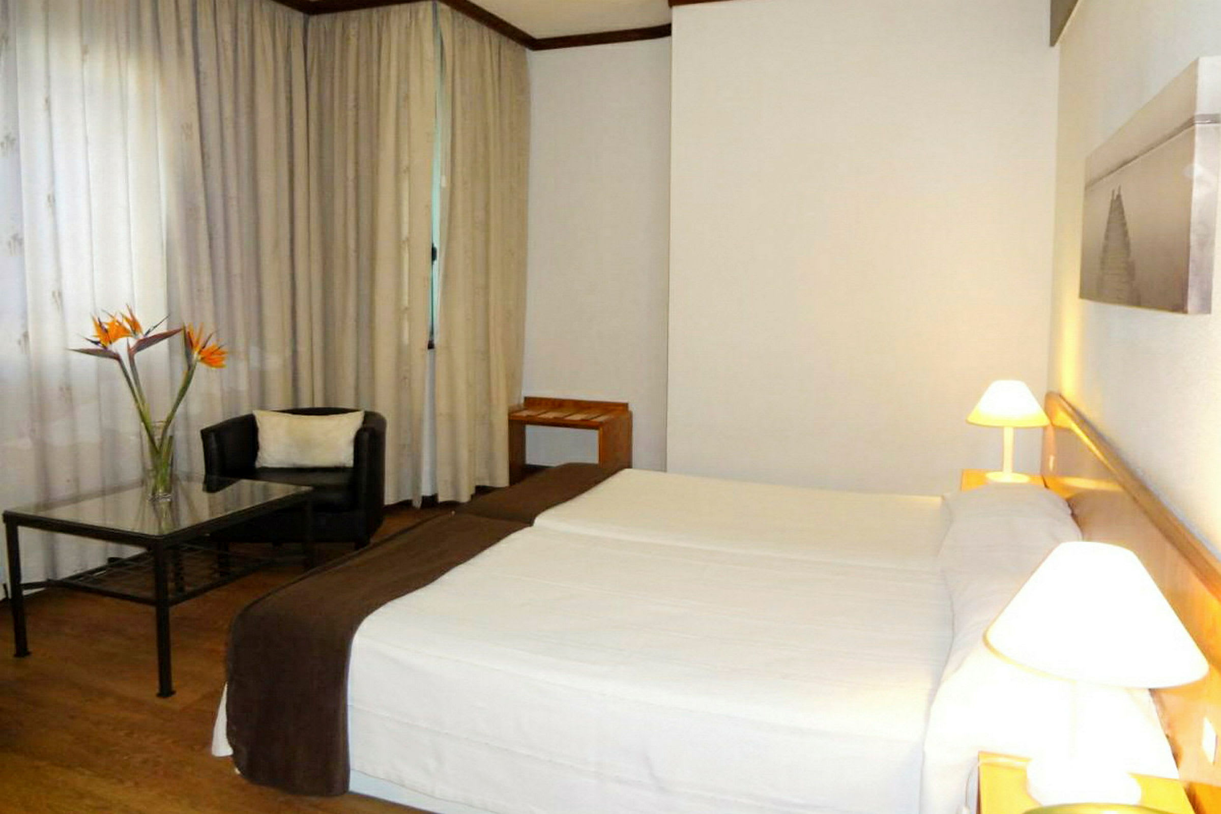 Hotellihuone, jossa on kaksi vuodetta, nojatuoli ja pöytä kukkamaljakolla. (Tekoälyn luoma teksti)