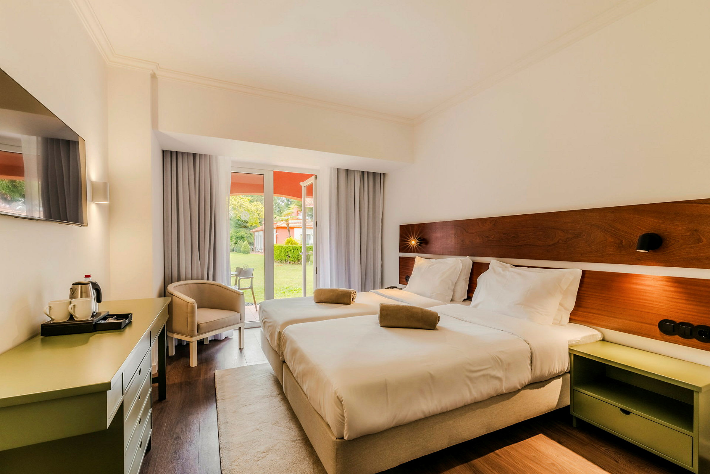Hotellihuone, jossa kaksi sänkyä, ja ikkunallinen ovi johtaa vehreään puutarhaan. (Tekoälyn luoma teksti)