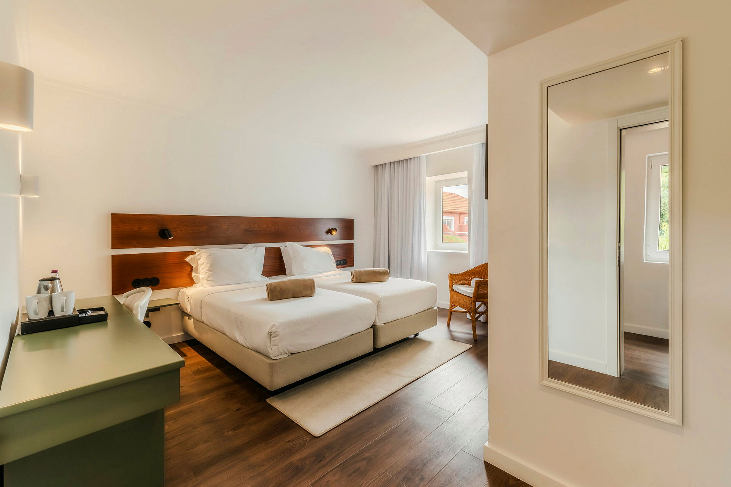 Hotellihuone, jossa on kaksi erillistä vuodetta, työpöytä ja seinäpeili. Huoneessa on suuret ikkunat verhoilla. (Tekoälyn luoma teksti)
