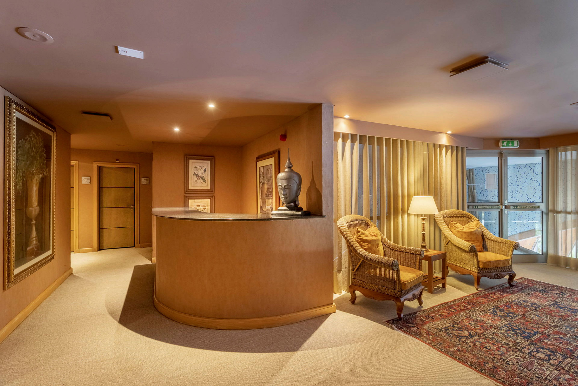 Sisäkuva kutsuvasta aulasta, jossa on kaksi nojatuolia ja lamppu, vastaanottotiski, taiteita seinillä ja matto. (Tekoälyn luoma teksti)