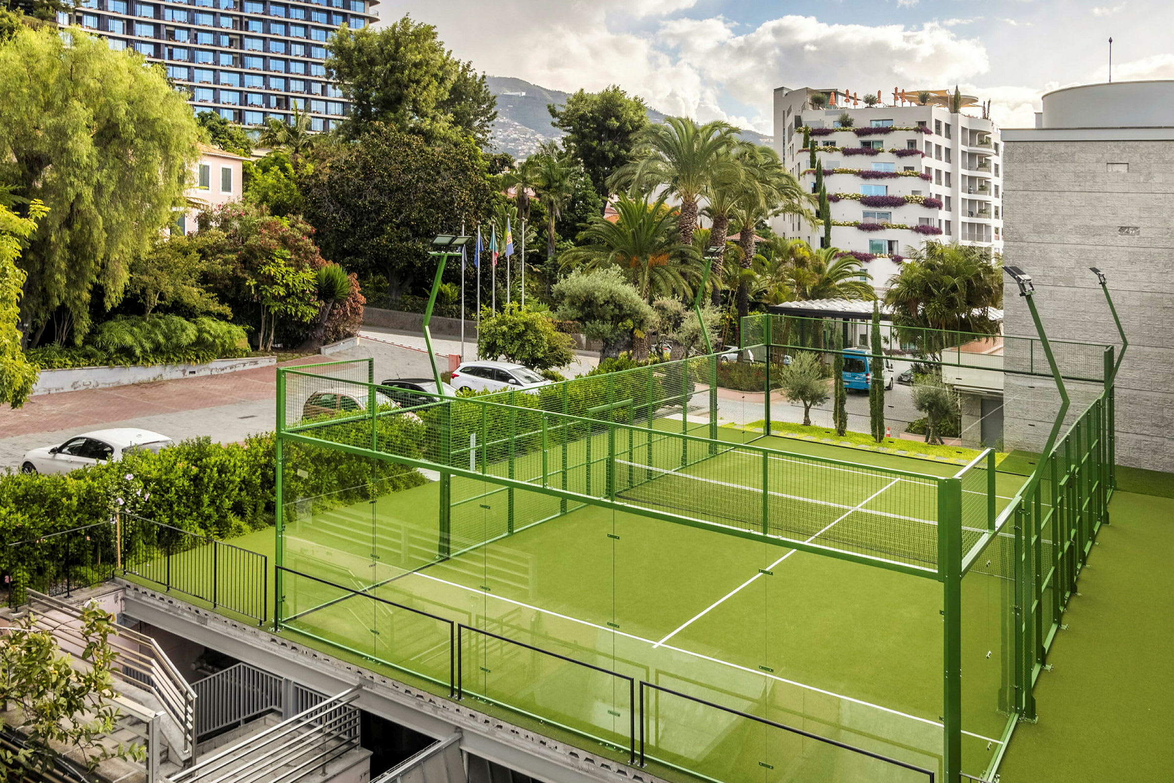 Vihreä padel-kenttä, jossa lasiseinät, puiden ja rakennusten ympäröimänä. (Tekoälyn luoma teksti)