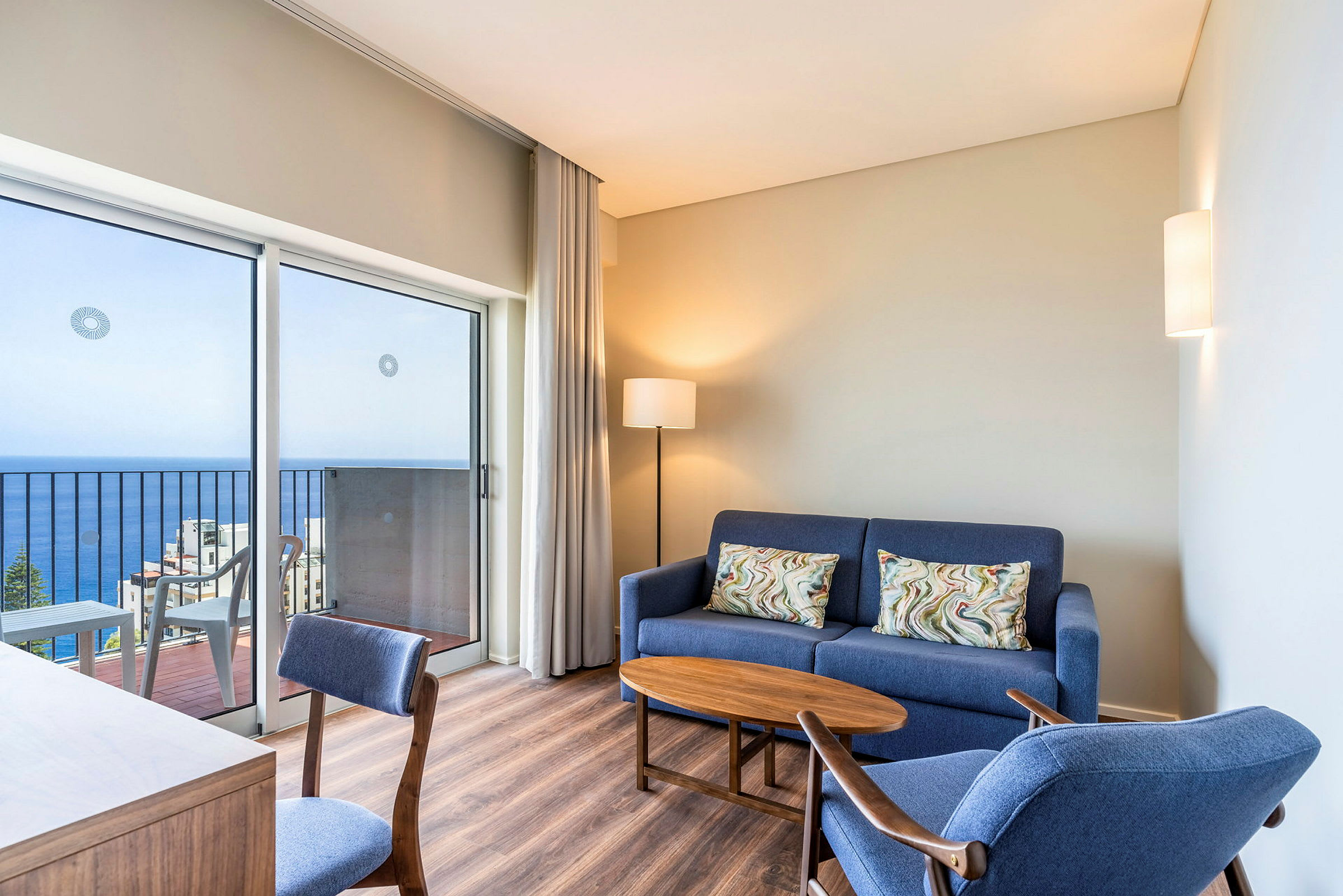 Modernisti sisustettu hotellihuone, jossa on sininen sohva, sininen nojatuoli, puupöytä ja työtuoli. Huoneessa on parveke merinäköalalla. (Tekoälyn luoma teksti)