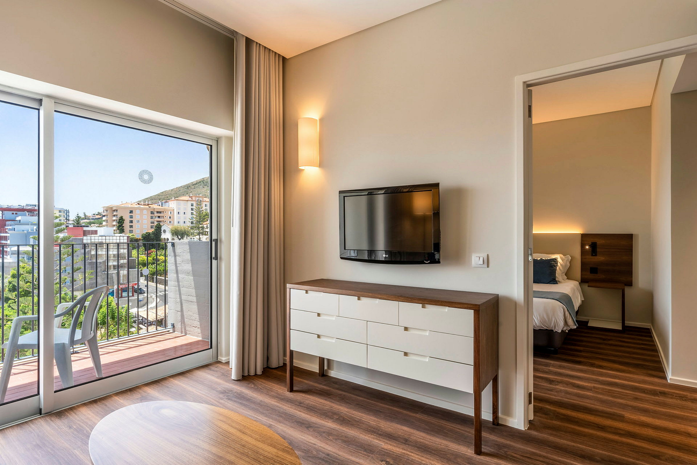 Hotellihuone, jossa seinällä televisio, lipasto ja viereisessä huoneessa sänky. Parvekkeelta avautuu näkymä rakennuksiin ja viheralueisiin. (Tekoälyn luoma teksti)