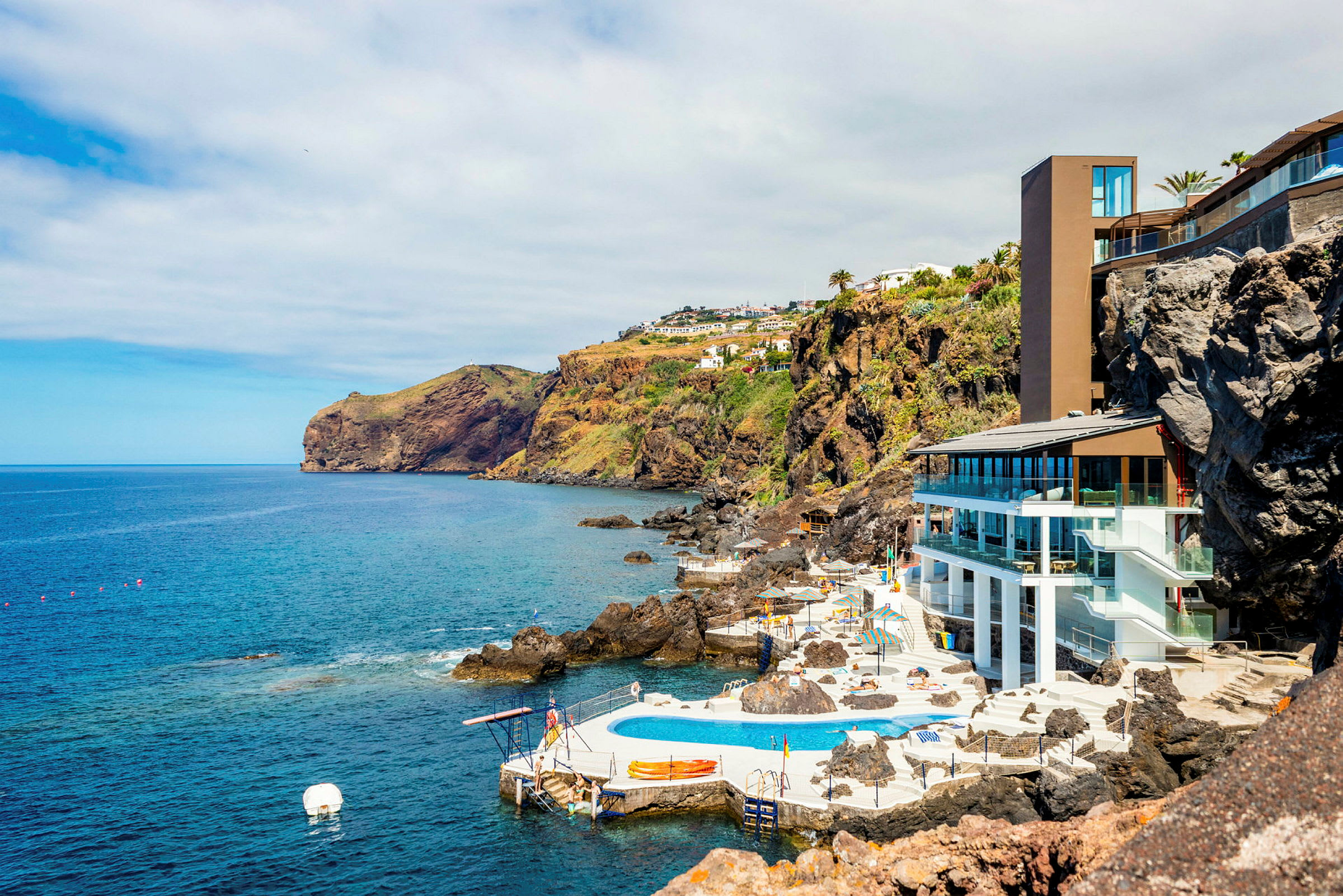 Rannikkomaisema, jossa on nykyaikainen rakennus ja uima-allasalue kallioiden laidalla, ympärillä meri ja vehreät rinteet. (Tekoälyn luoma teksti)