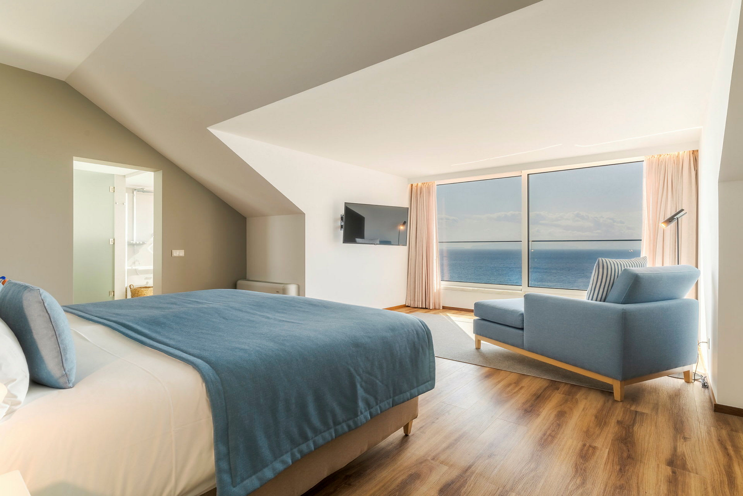 Moderni hotellihuone merinäköalalla, sininen päiväpeite sängyssä ja sininen sohva. (Tekoälyn luoma teksti)