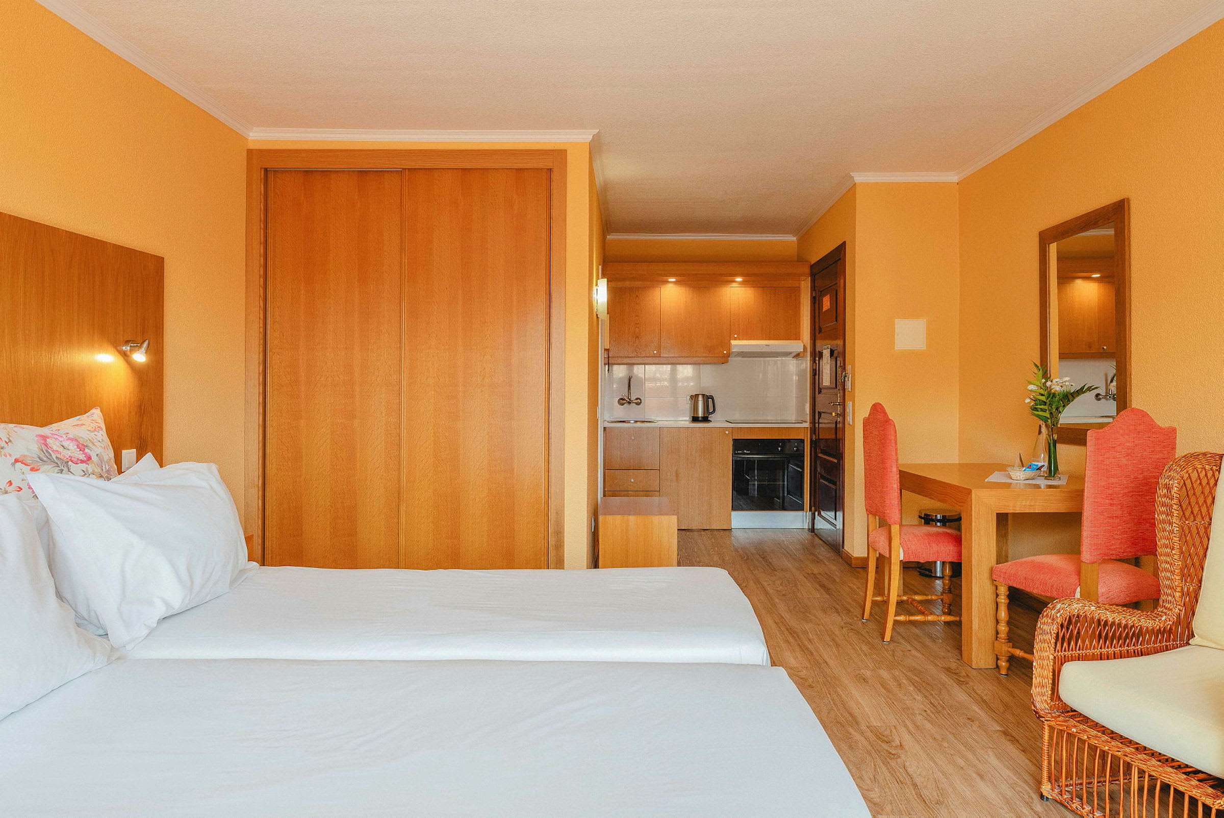 Hotellihuone kahdella erillisellä sängyllä, puukalusteilla, oransseilla seinillä ja pienellä keittiöllä taustalla. (Tekoälyn luoma teksti)