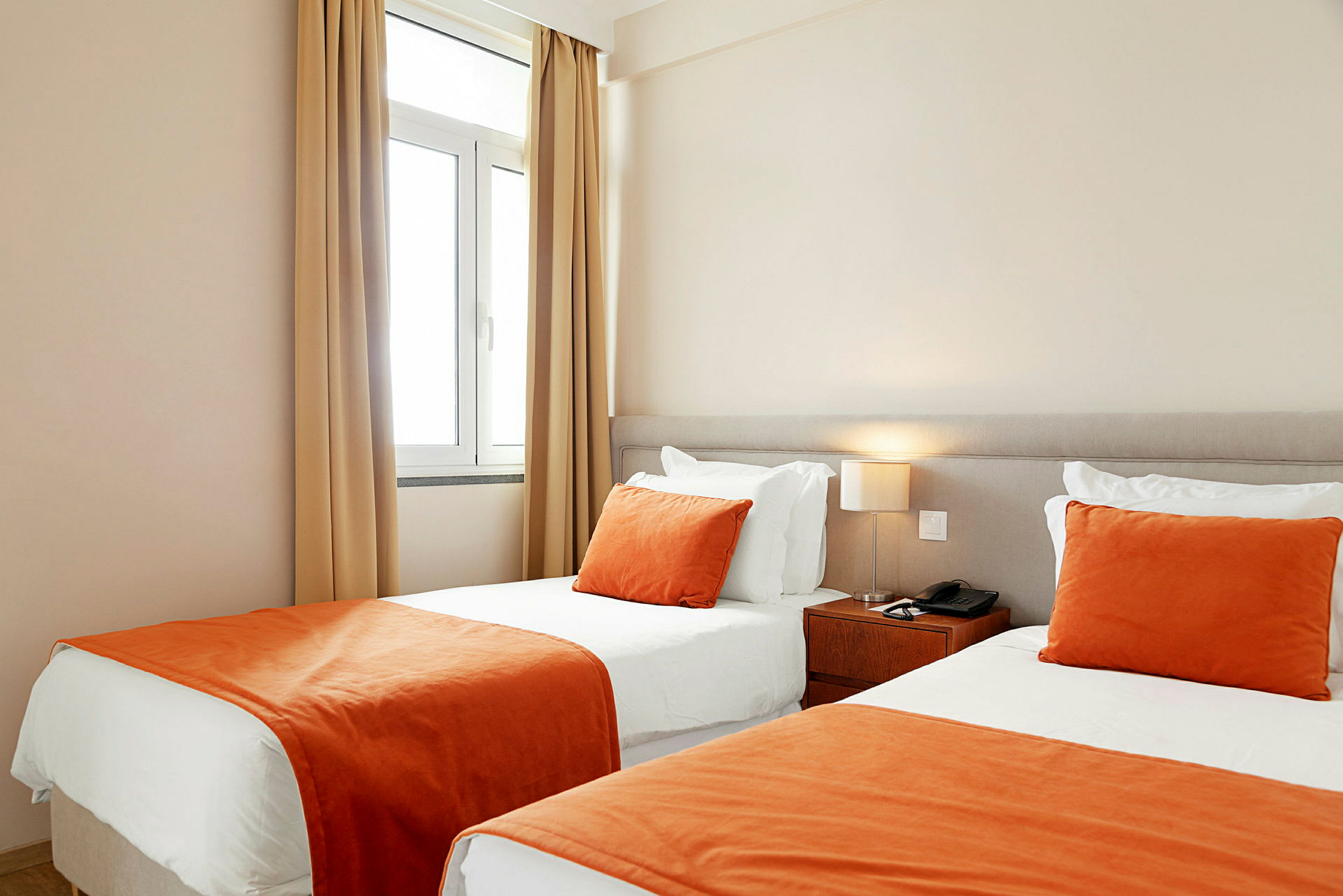 Hotellihuone, jossa on kaksi vierekkäistä yhden hengen vuodetta, valkoiset lakanat ja oranssit päiväpeitteet sekä koristetyynyt. (Tekoälyn luoma teksti)