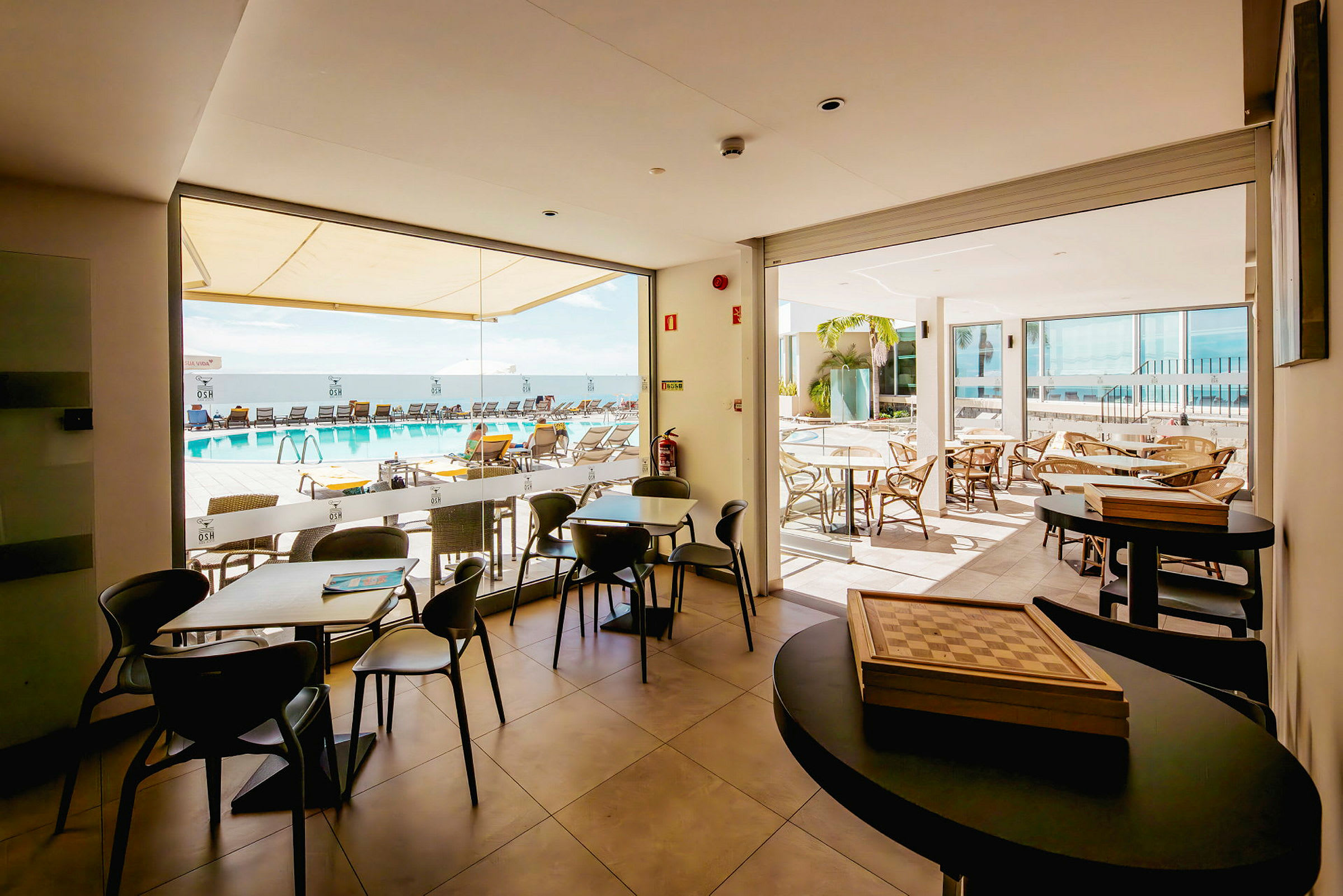Modernin kahvilan sisätila, jossa lasiseinät ja näkymä ulkoalueelle, jossa on uima-allas ja aurinkotuoleja. (Tekoälyn luoma teksti)