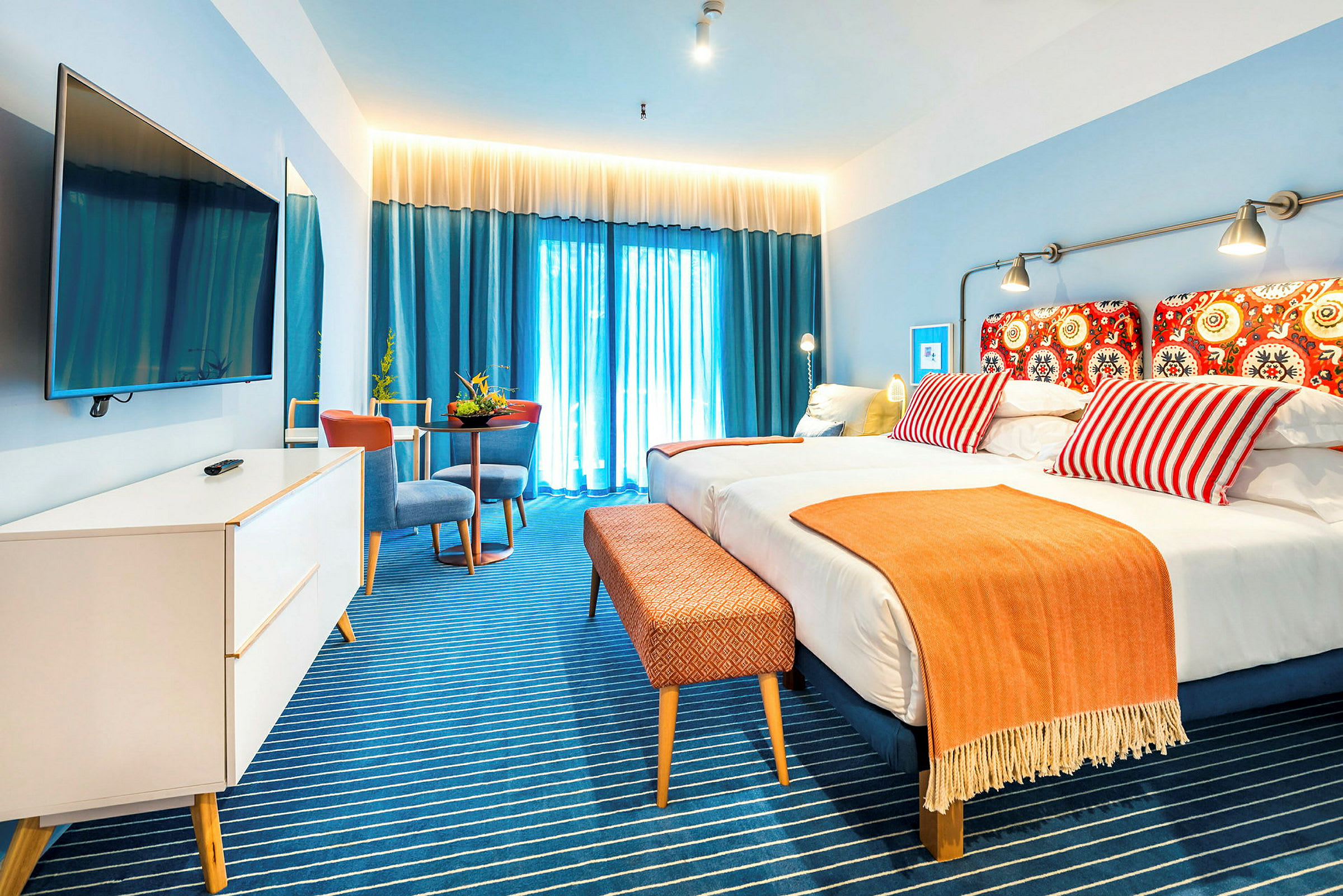 Hotellihuone, jossa parivuode, värikkäät tyynyt ja oranssi huopa. Siniset verhot ja valon sisäänkäynti taustalla. (Tekoälyn luoma teksti)