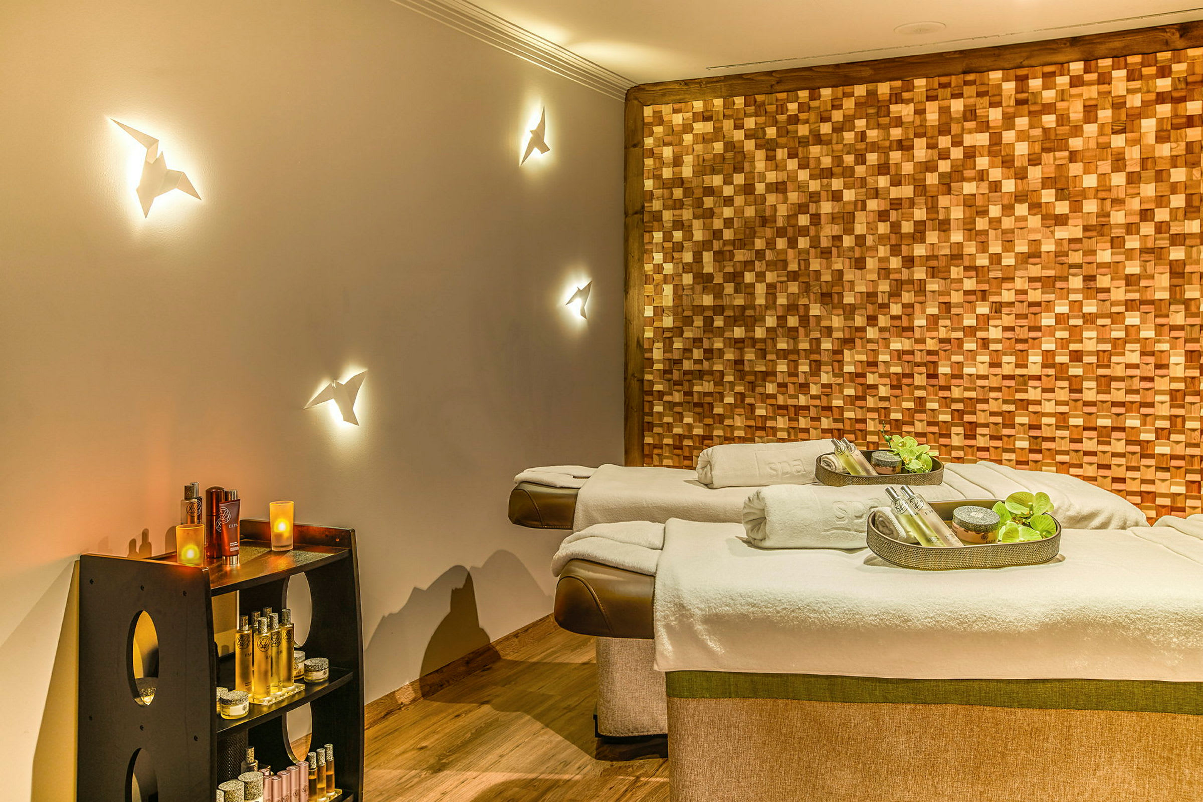 Kaksi spa-hierontapöytää koristeellisilla lisävarusteilla, ympäröity puupaneeliseinällä ja hyllyillä, joissa on erilaisia kylpylätuotteita. (Tekoälyn luoma teksti)