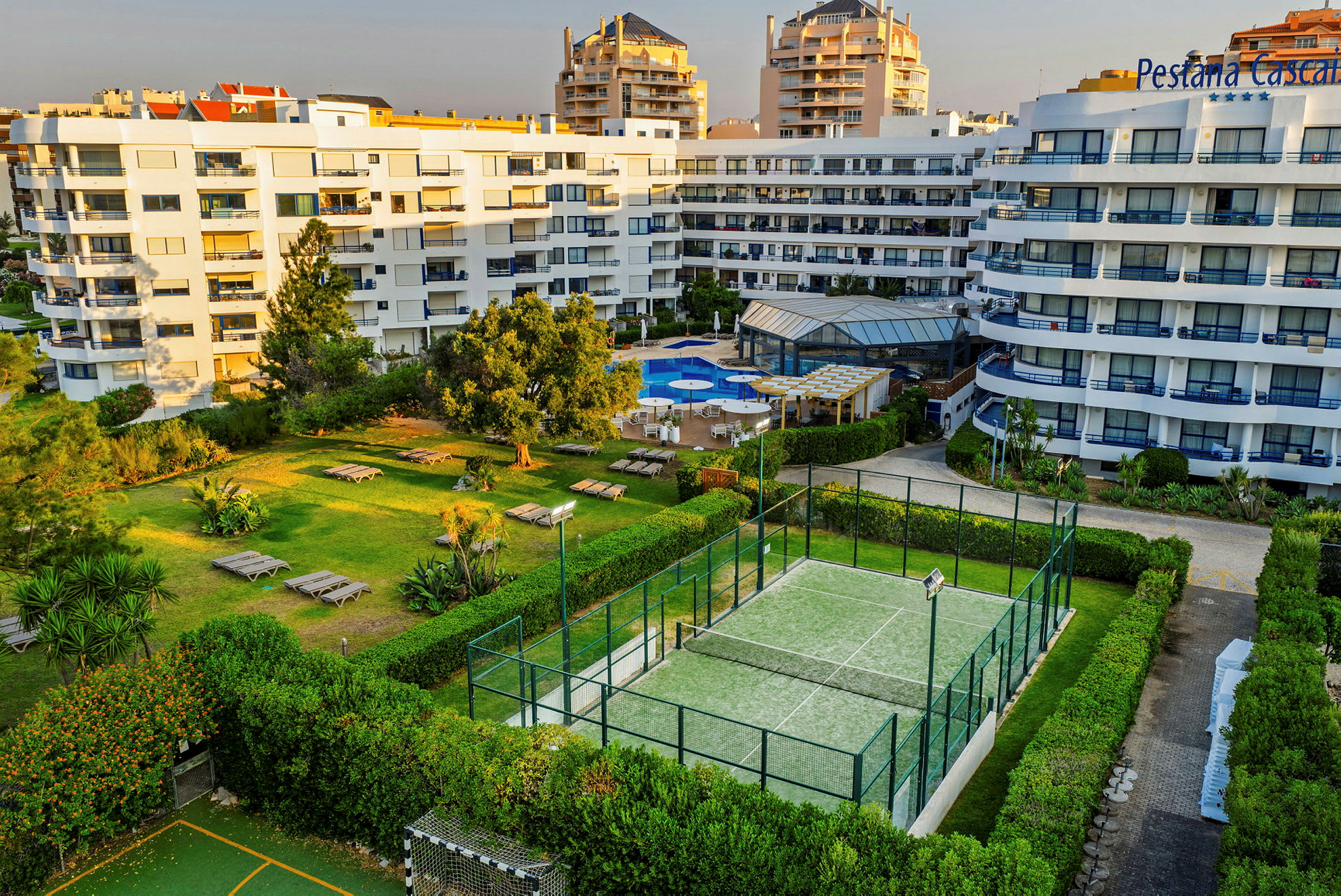 Valkopintainen rakennus, uima-allasalue aurinkotuoleineen ja pergoloineen sekä tenniskenttä, ympäröity vihreillä alueilla ja pensailla. (Tekoälyn luoma teksti)
