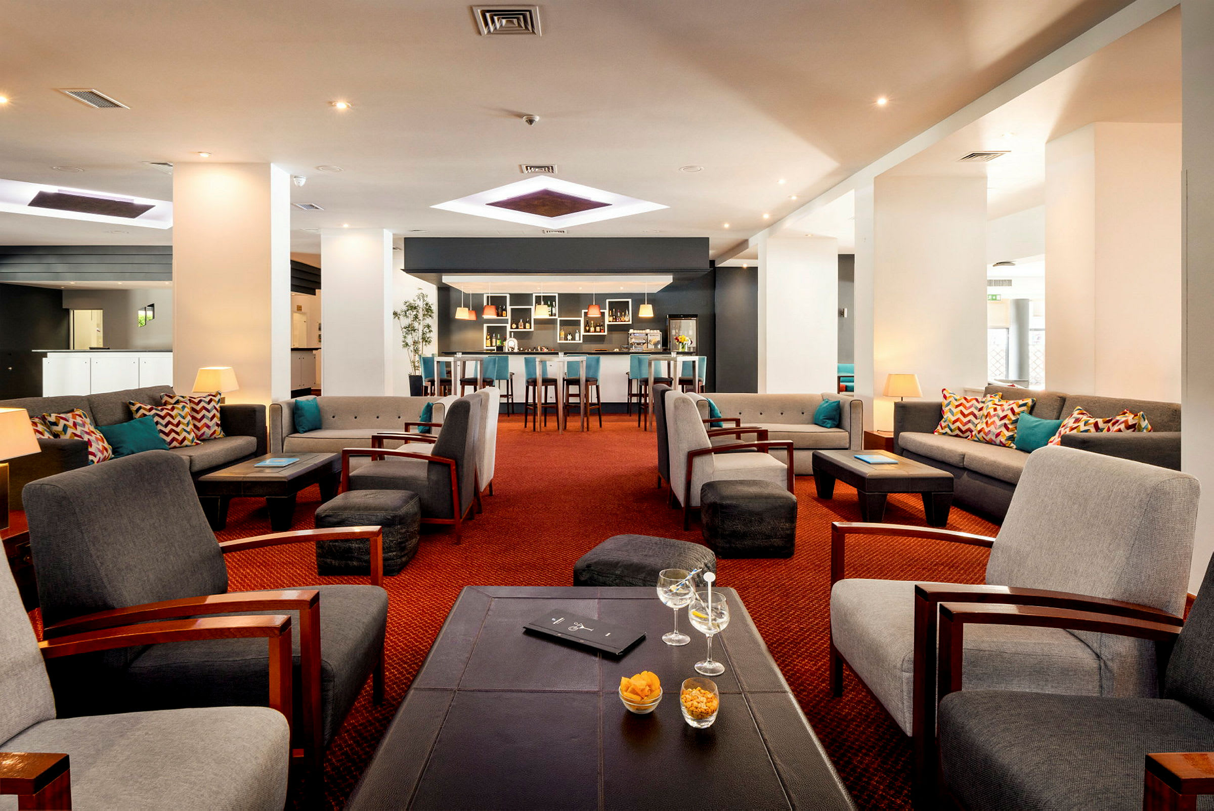 Lounge, jossa mukavia istumapaikkoja, sohvia ja nojatuoleja pienten pöytien ympärillä. Seinät ovat valkoiset, ja taustalla näkyy baari. (Tekoälyn luoma teksti)