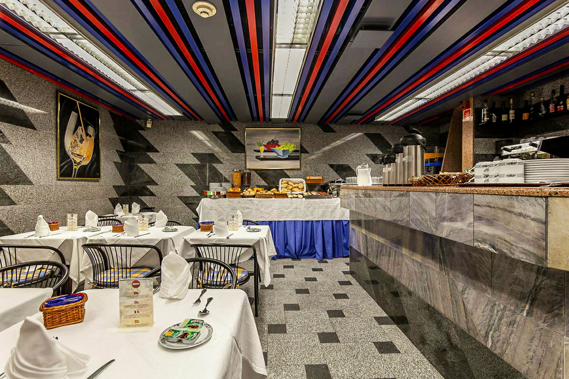 Huone, jossa on katetut pöydät, tuolit ja buffet erilaisilla ruoka-aineilla. Seinät ja lattia on kuvioitu geometrisilla kuvioilla. (Tekoälyn luoma teksti)