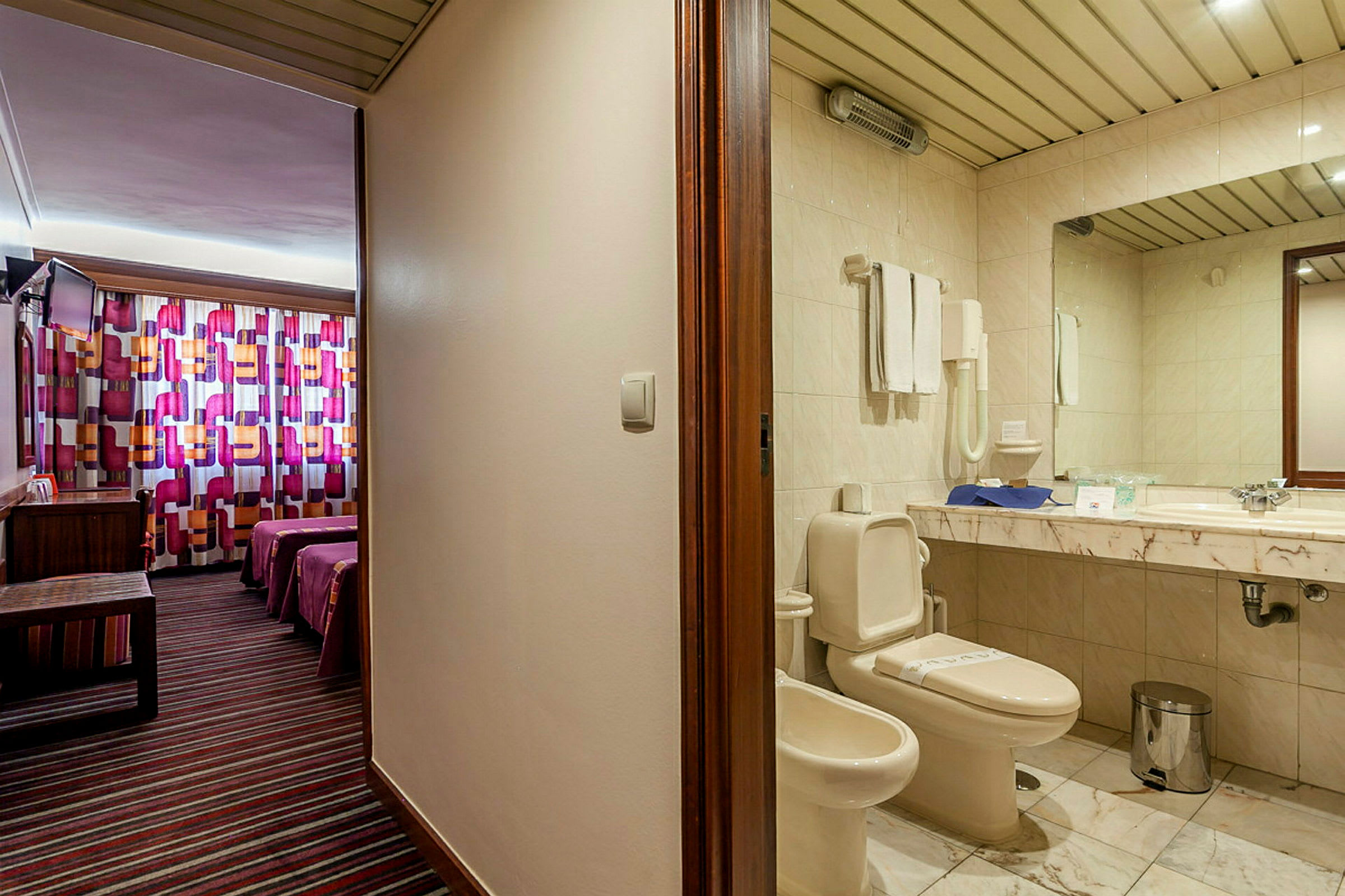 Hotellihuoneen sisätilat, jossa vasemmalla on kylpyhuone, jossa wc ja pesuallas, ja oikealla värikkäillä verhoilla varustettu makuuhuone kahdella sängyllä. (Tekoälyn luoma teksti)