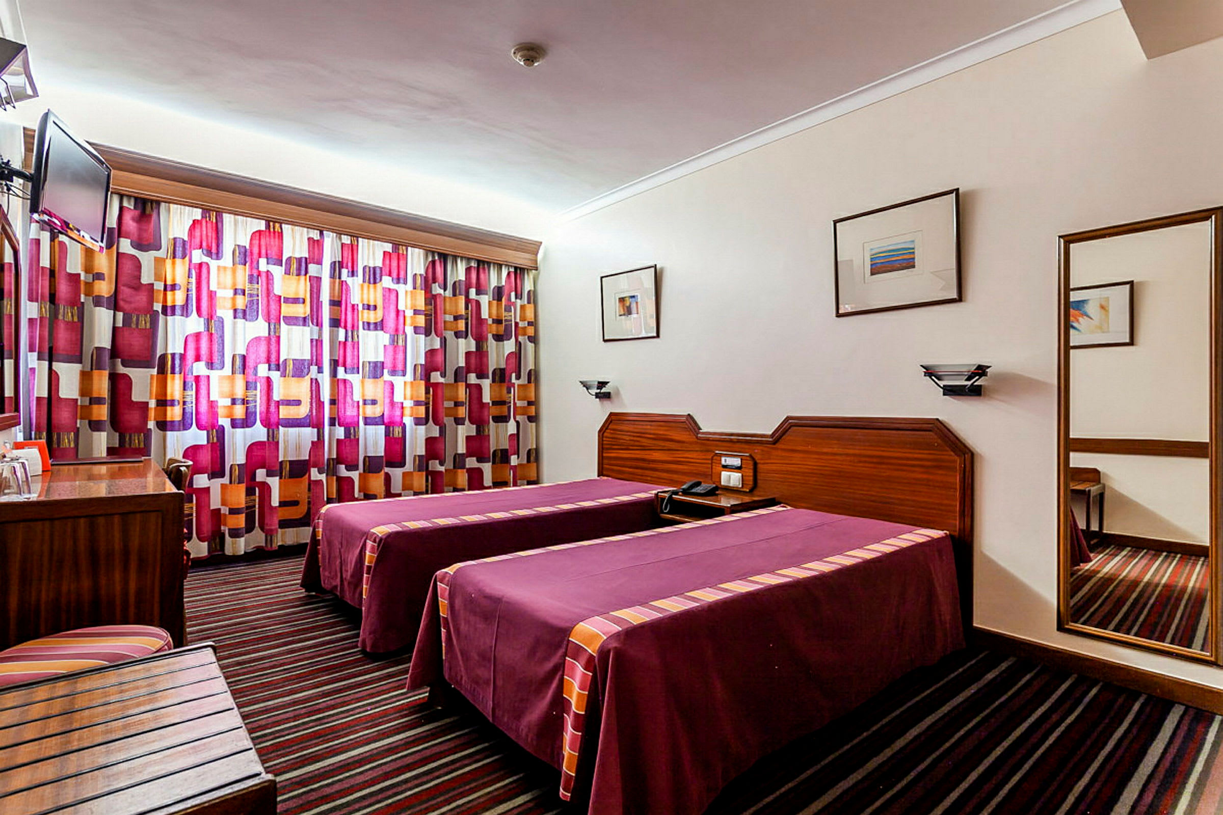 Hotellihuone kahdella yhden hengen vuoteella, värikkäillä verhoilla ja seinään asennetulla peilillä. (Tekoälyn luoma teksti)