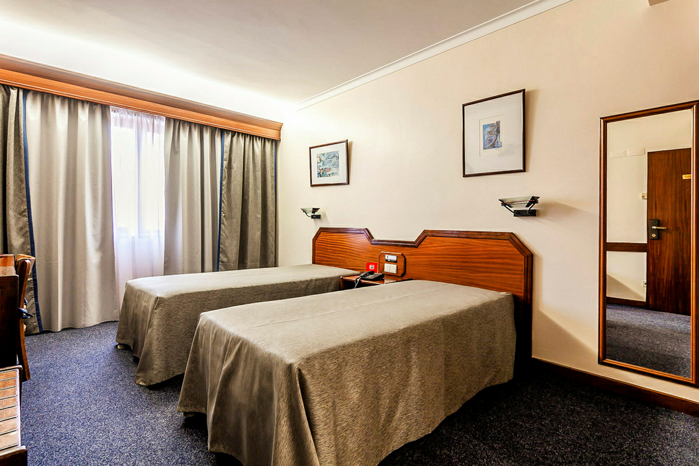 Kuva hotellihuoneesta, jossa on kaksi yhden hengen vuodetta, työpöytä ja ikkuna verhoineen. (Tekoälyn luoma teksti)