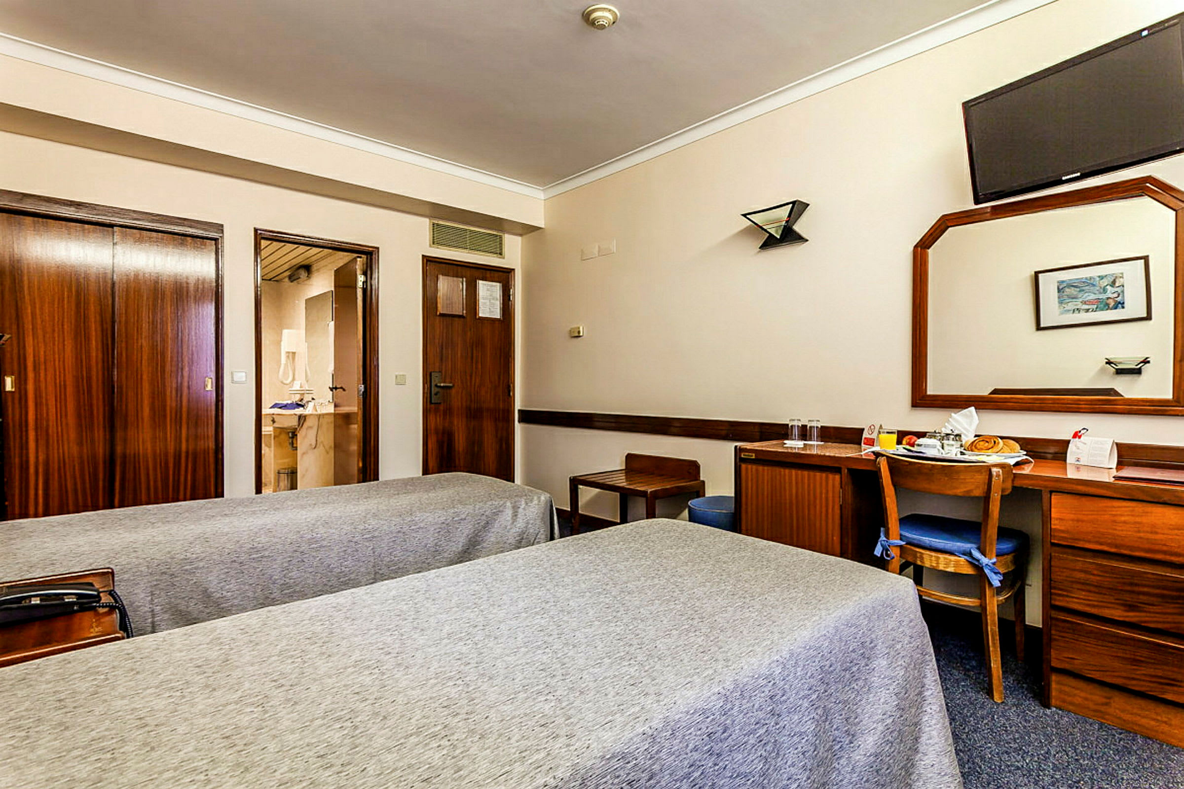 Hotellihuone, jossa on kaksi yhden hengen vuodetta, puinen kirjoituspöytä tuoleineen, suuri peili ja seinään asennettu televisio. (Tekoälyn luoma teksti)